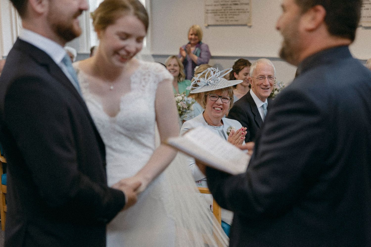 Joyful wedding ceremony with bride in lace gown, guests smiling, woman in floral hat laughing in church.