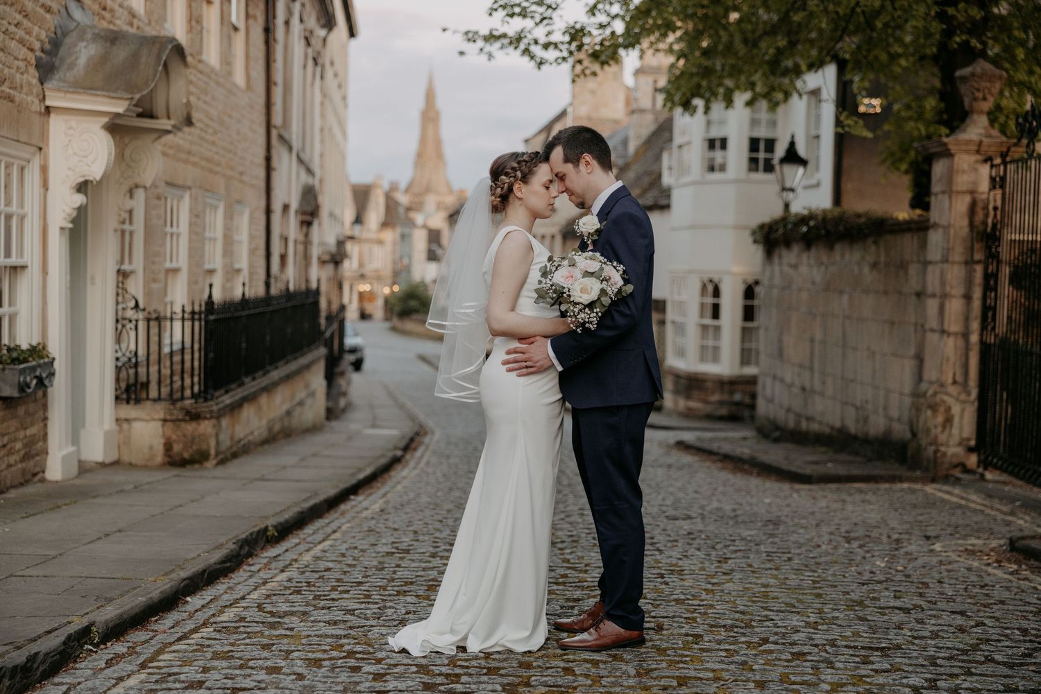 Bride and groom share romantic moment on cobblestone street, bride in white gown, groom in navy suit, holding bouquet.