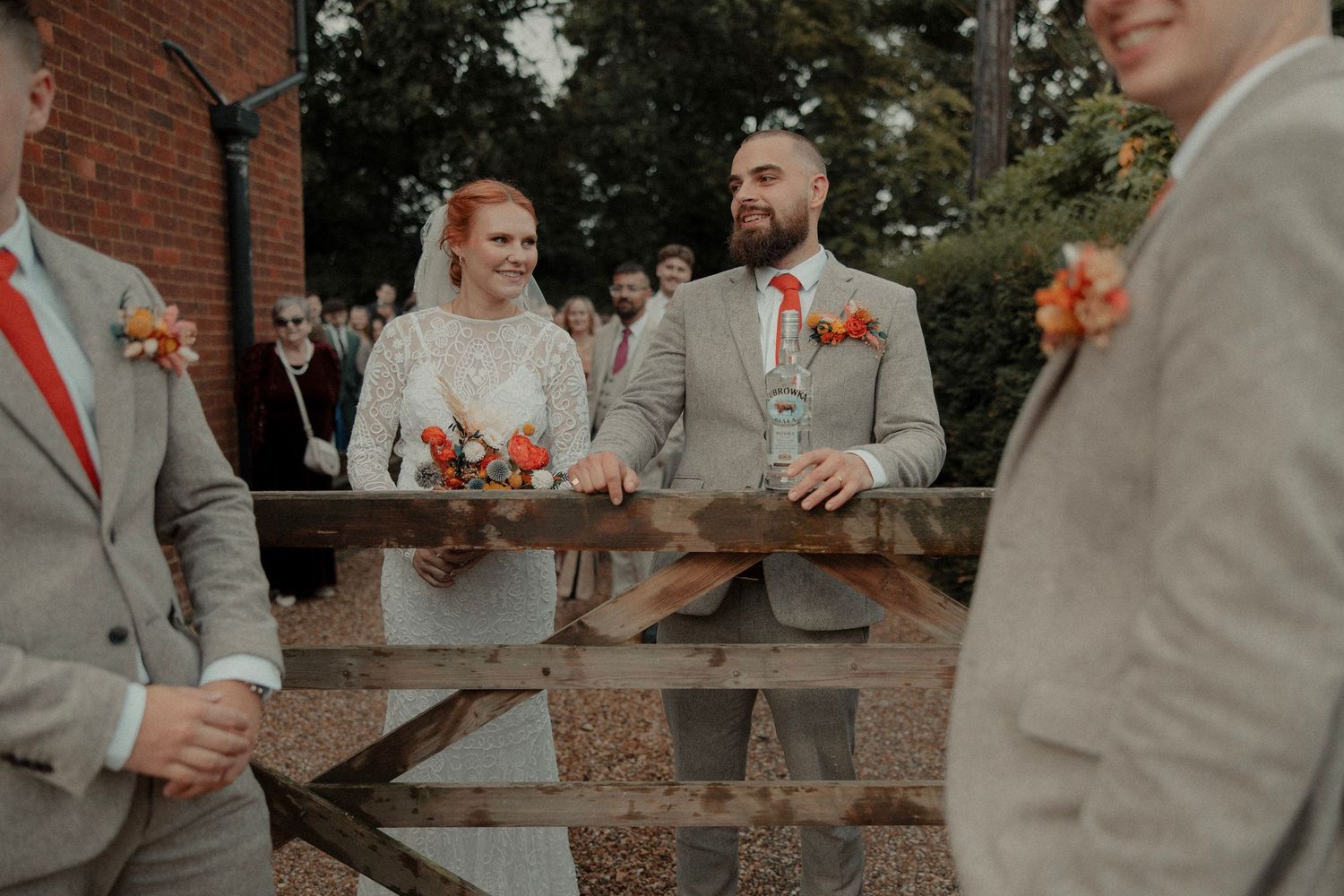 Bride and groom smile at wedding ceremony outdoors, flanked by groomsmen in grey suits with orange boutonnieres.