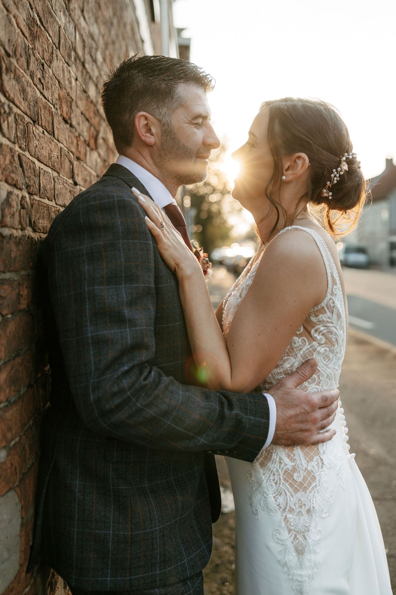 A bride and groom share a romantic golden hour moment leaning against a brick wall, with the sun glowing warmly behind them.