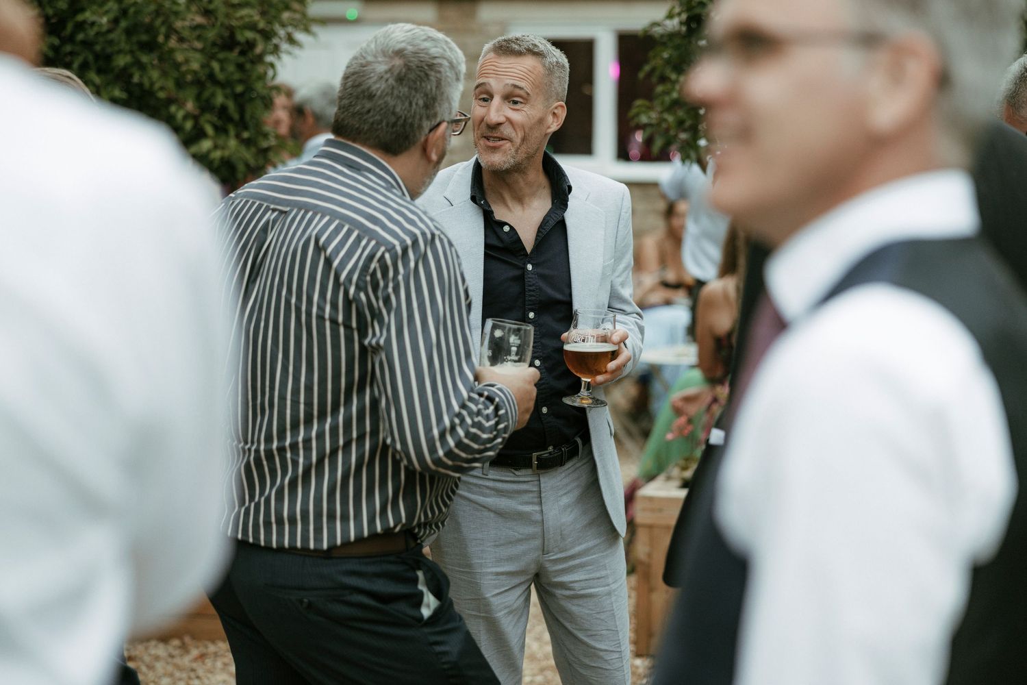 Men in smart casual attire socializing outdoors at a garden party, holding drinks and engaging in conversation.