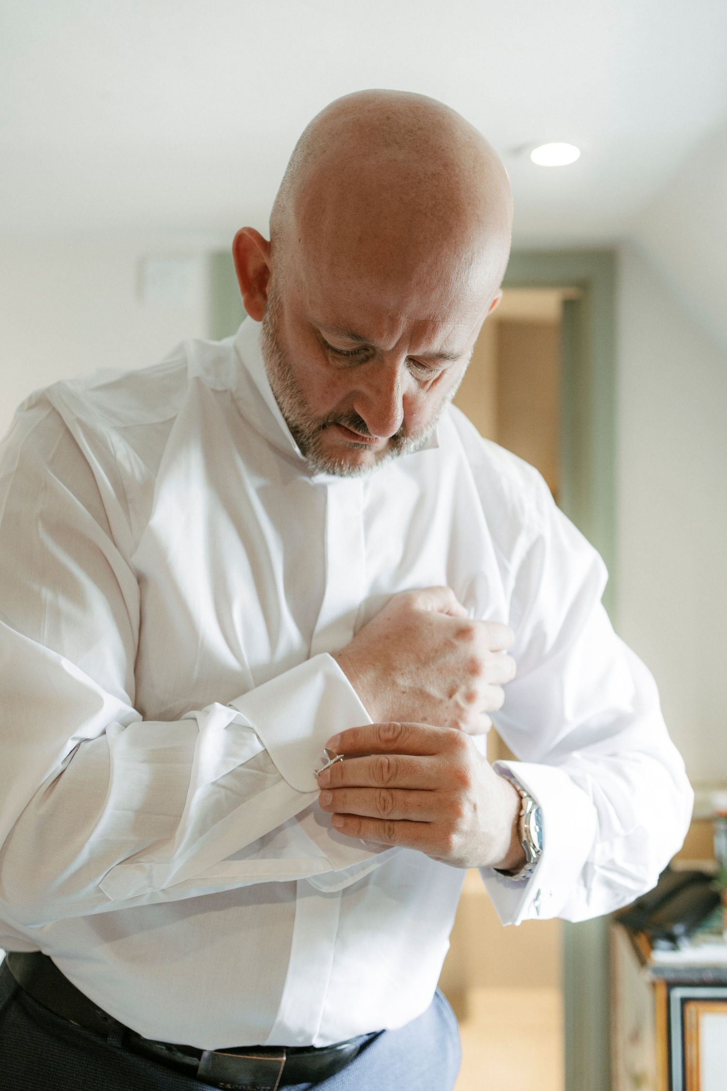 Bald groom in white dress shirt fastening cufflinks while getting ready for his wedding day.