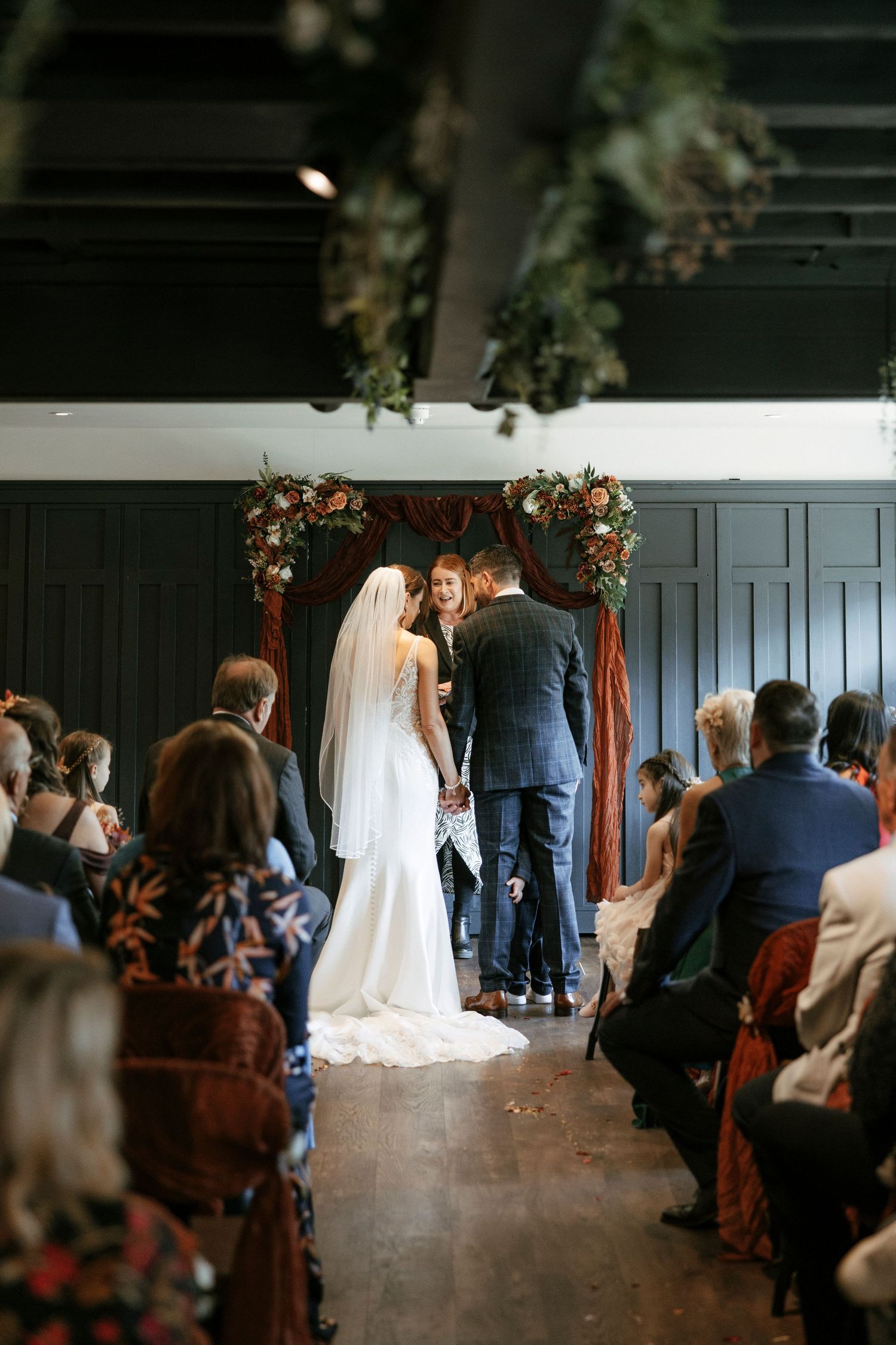 Bride and groom exchange vows at floral arch altar surrounded by seated wedding guests indoors.