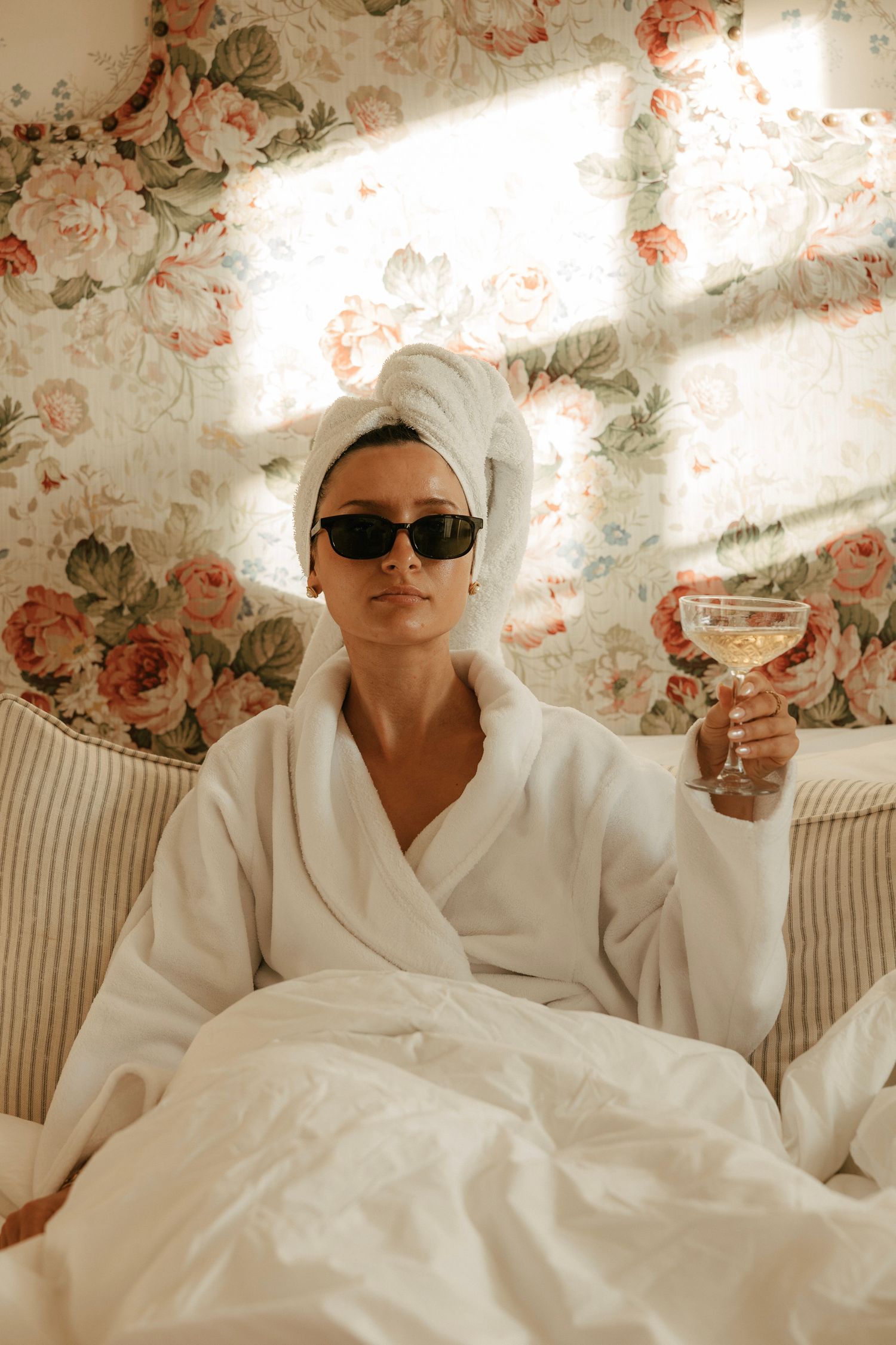 Woman in white bathrobe and towel turban wearing sunglasses, holding a cocktail glass in bed with floral wallpaper.