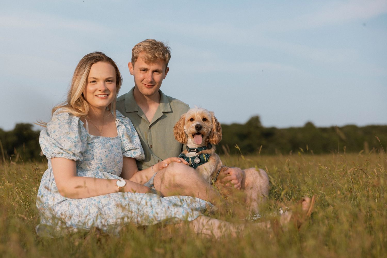 Couple sitting in a sunny green field with a golden cocker spaniel dog, smiling outdoors on a clear day.