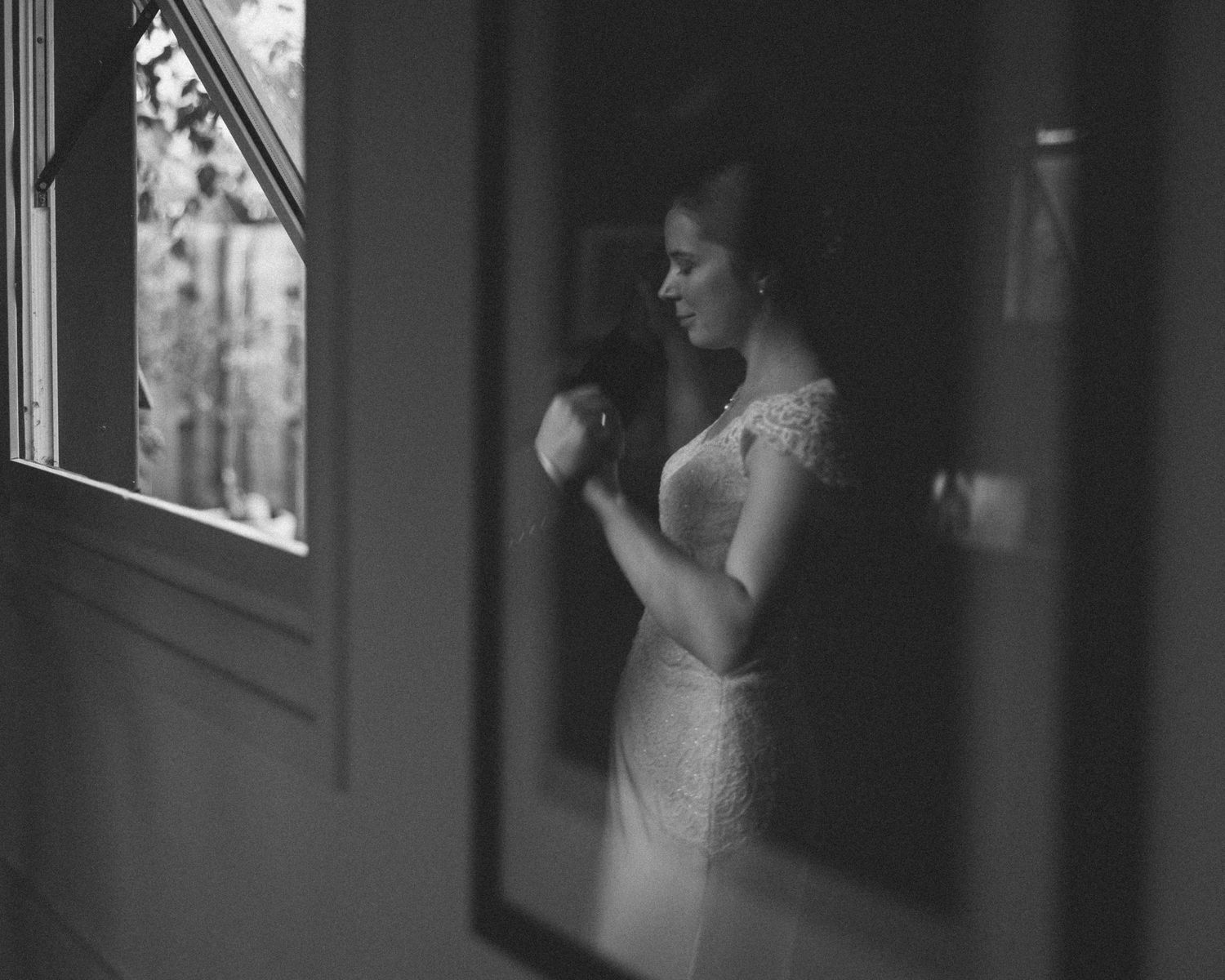 Black and white photo of a bride in lace wedding dress standing by a window, adjusting her gown in soft natural light.