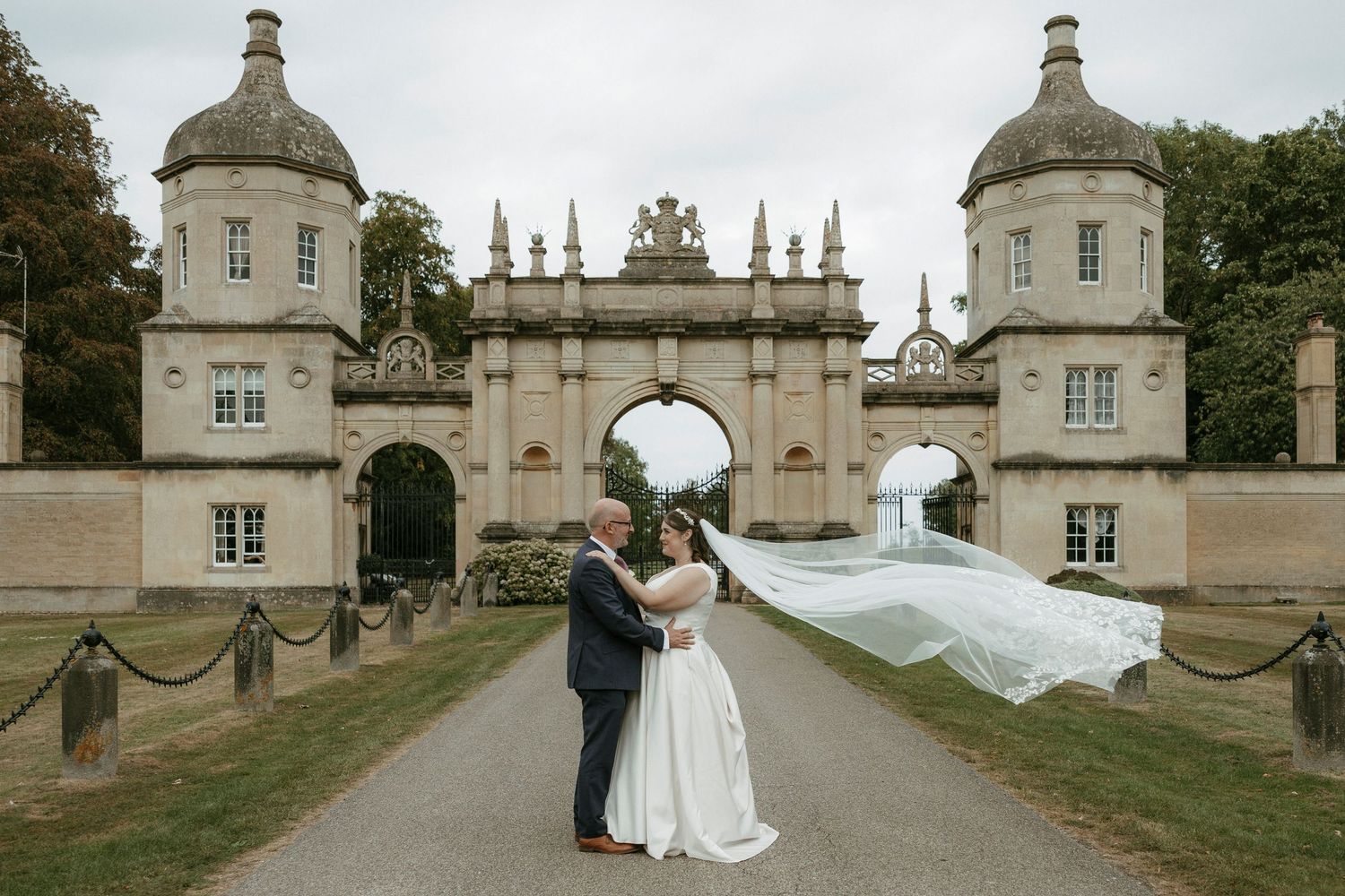 Bride and groom embrace before grand stone gatehouse as wedding veil flows dramatically in the wind.