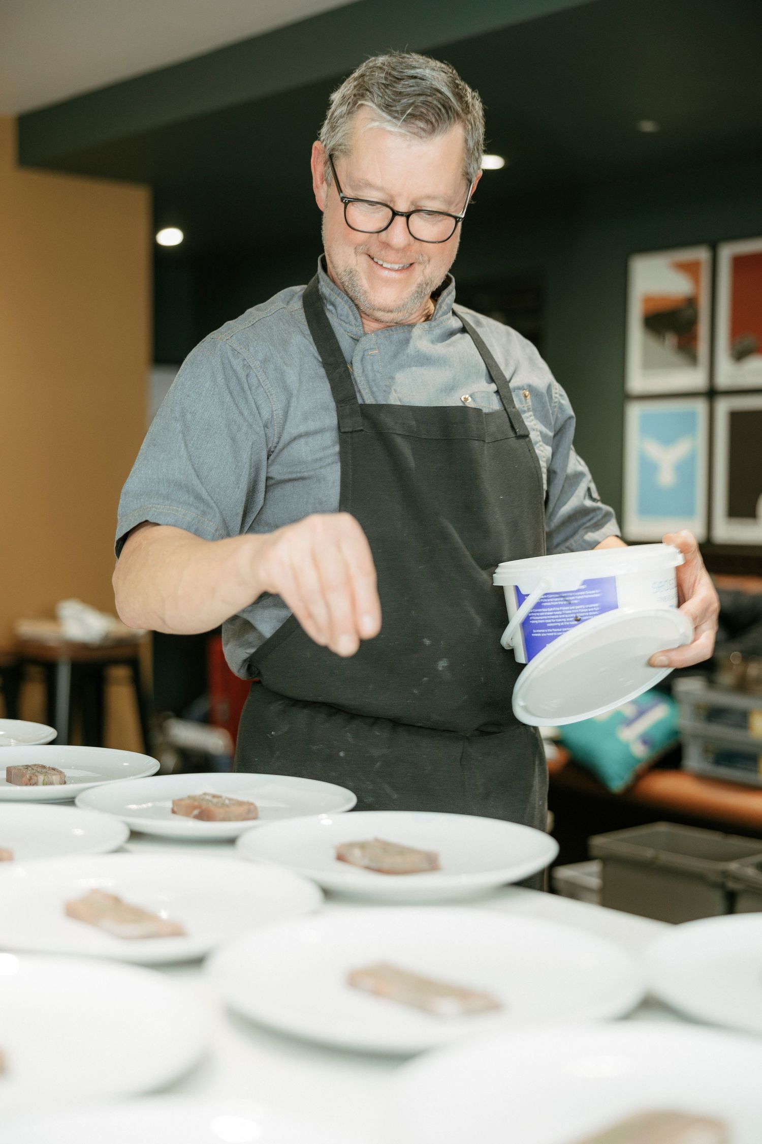 Professional chef in glasses and black apron preparing plated dishes in a restaurant kitchen.