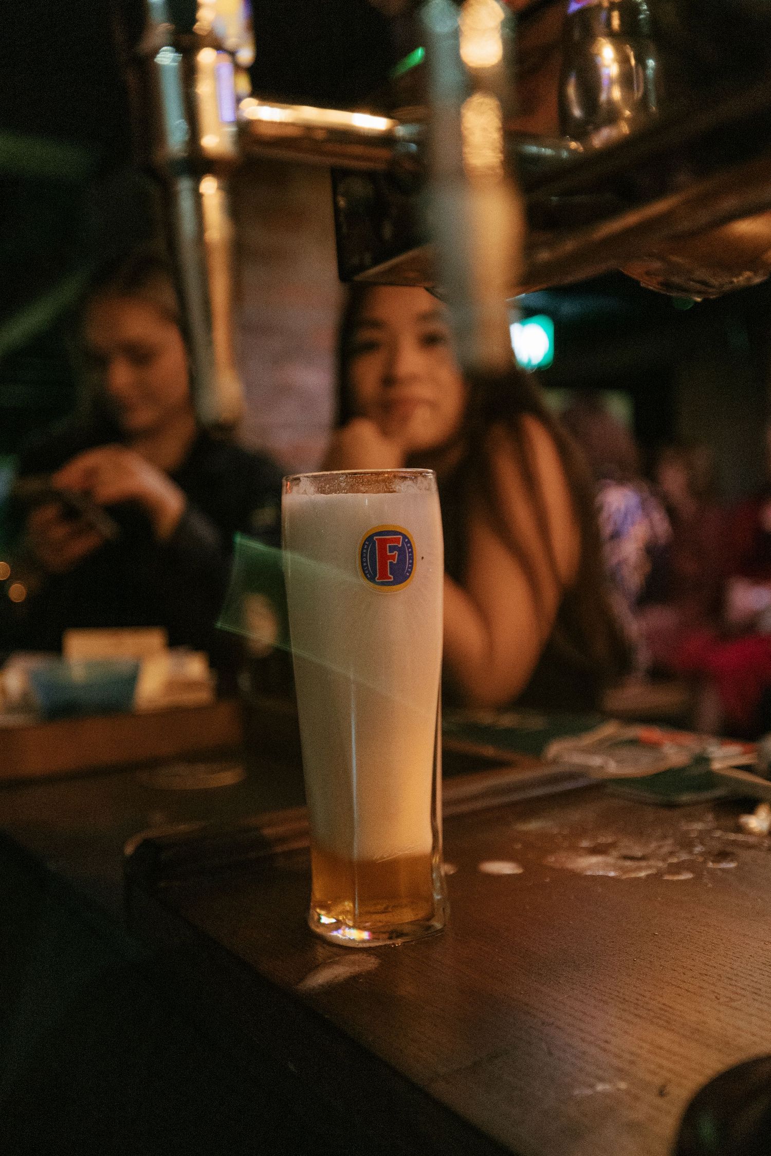 Tall glass of light beer with foam head sits on dark wooden bar counter in moody lighting.