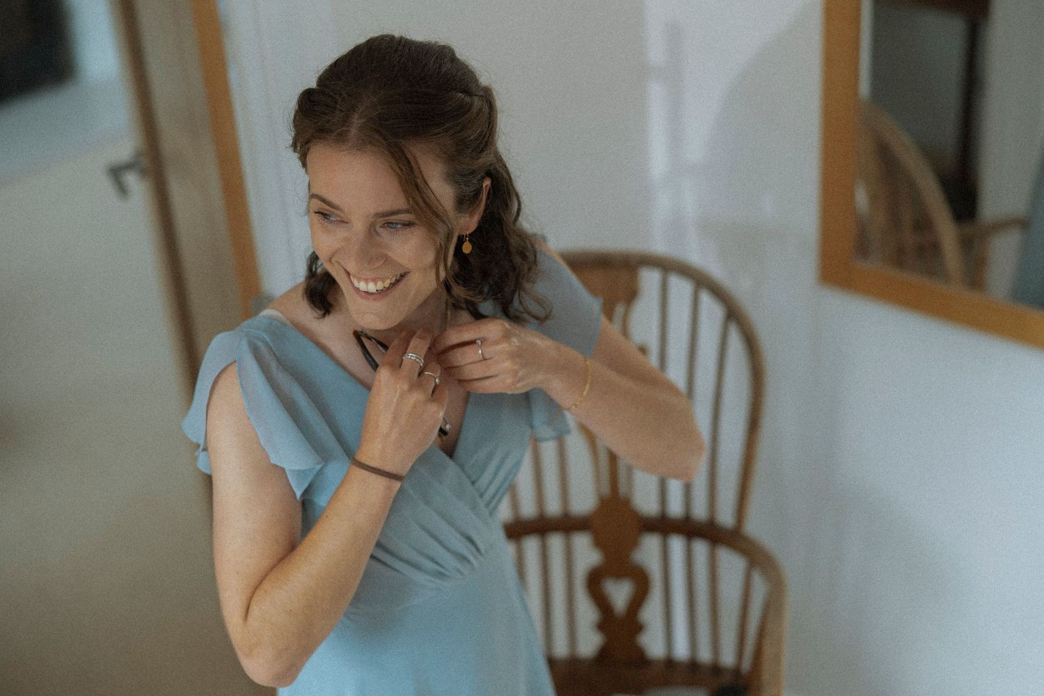 Smiling woman in blue dress fastening necklace near wooden chair in bright room.