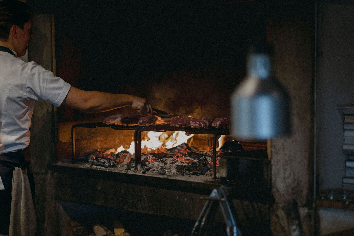 Chef grilling meat over open wood fire in a rustic stone hearth restaurant kitchen.