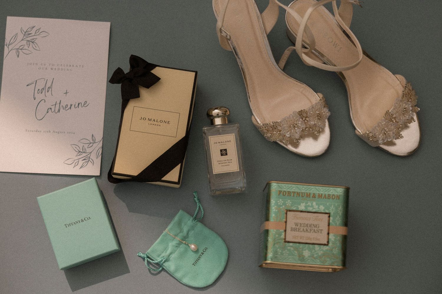Elegant bridal flat lay featuring Jo Malone perfume, Tiffany jewelry, embellished heels, and wedding invitation on gray background.