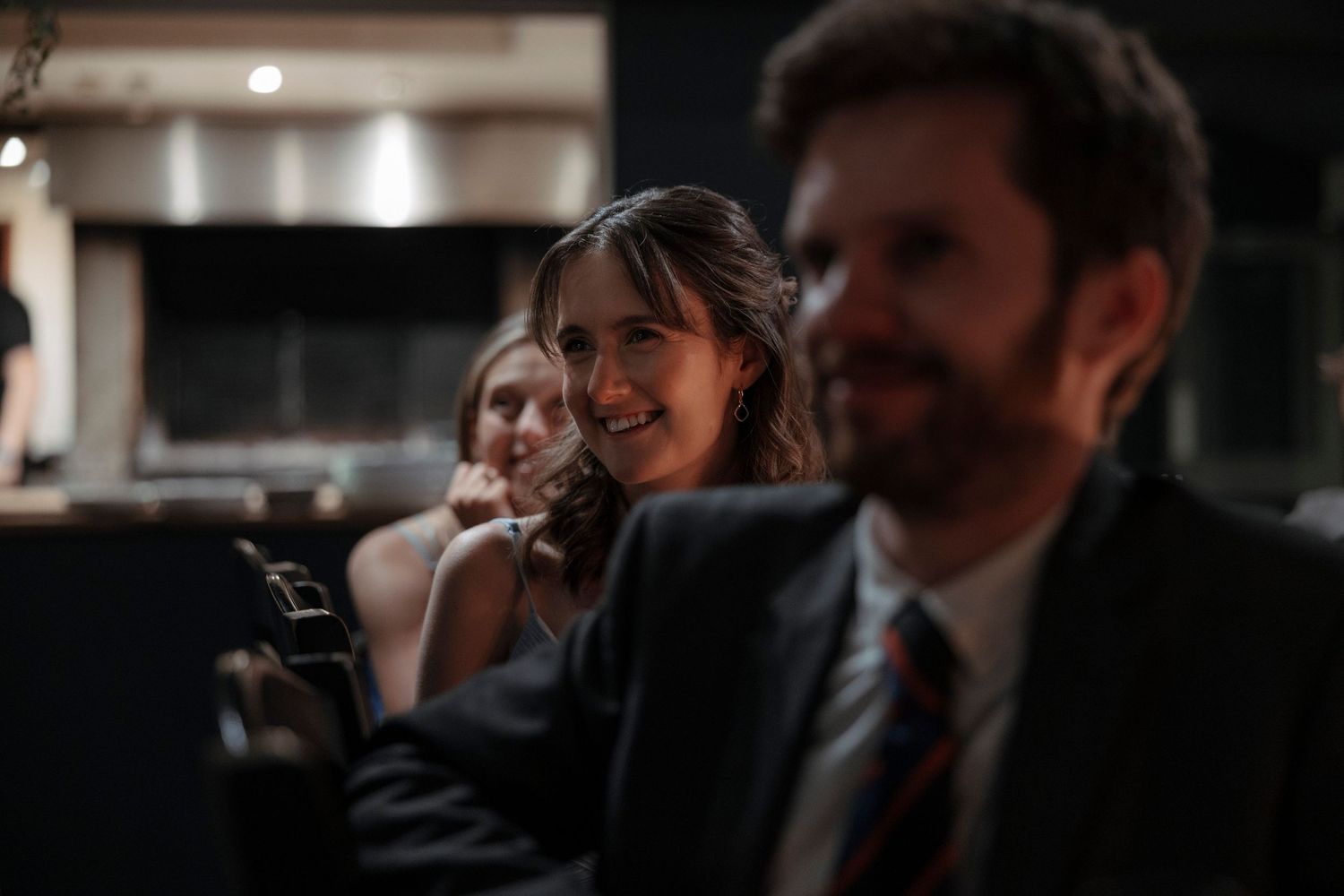 A smiling woman in formal attire sits between guests at an elegant indoor event, with a bearded man in a suit visible in the foreground.