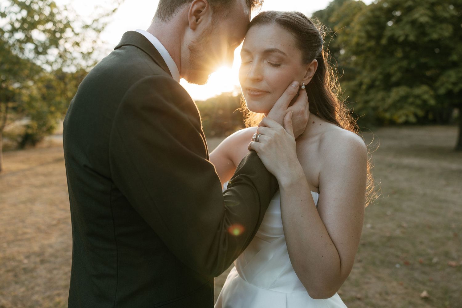 A groom tenderly holds his bride's face at golden hour, sunlight glowing behind them in a romantic outdoor wedding portrait.