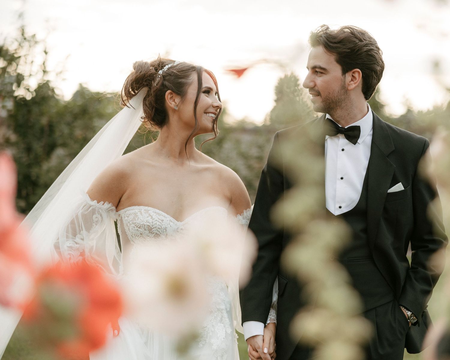 A bride and groom hold hands outdoors, smiling at each other, she in a white gown and veil, he in a black tuxedo.