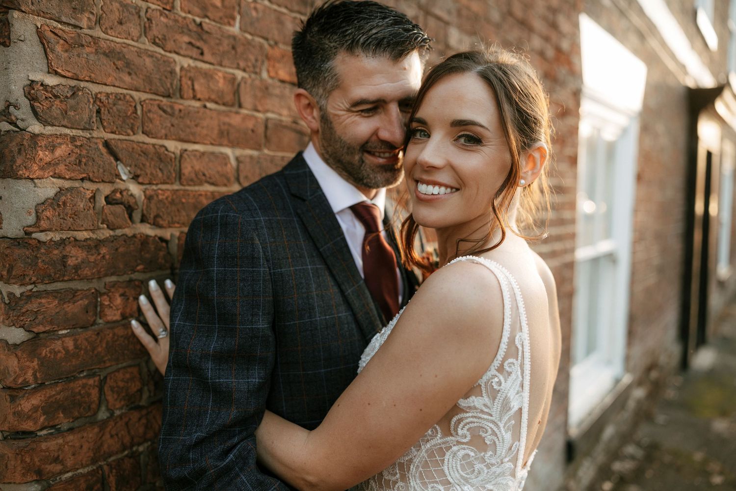 A smiling bride in a lace gown embraces a groom in a plaid suit against a rustic brick wall.