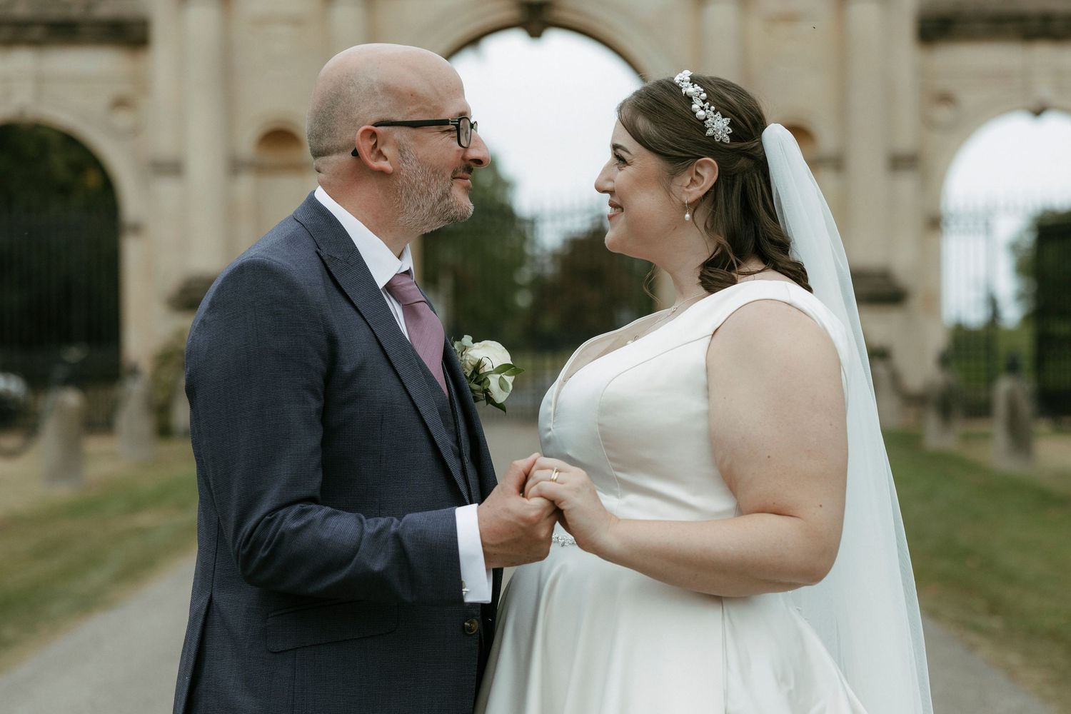 Bride and groom holding hands and smiling at each other outdoors near an ornate stone arch on their wedding day.