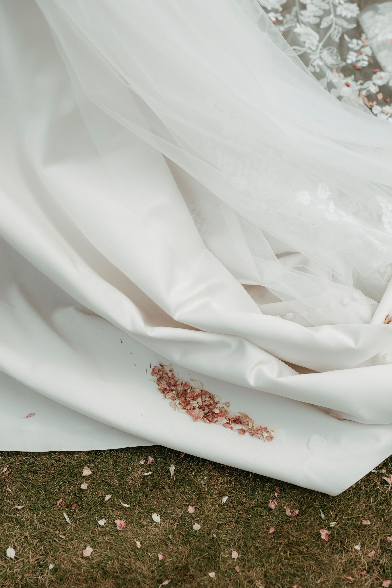 White wedding dress train spread on grass with pink confetti petals scattered on the fabric.