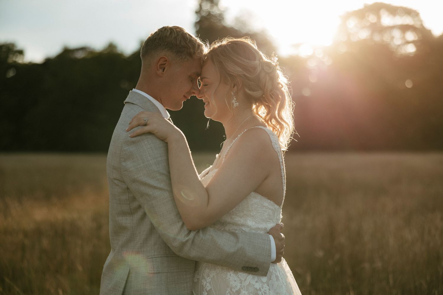A couple embraces in a golden sunlit field during sunset while wearing wedding attire.