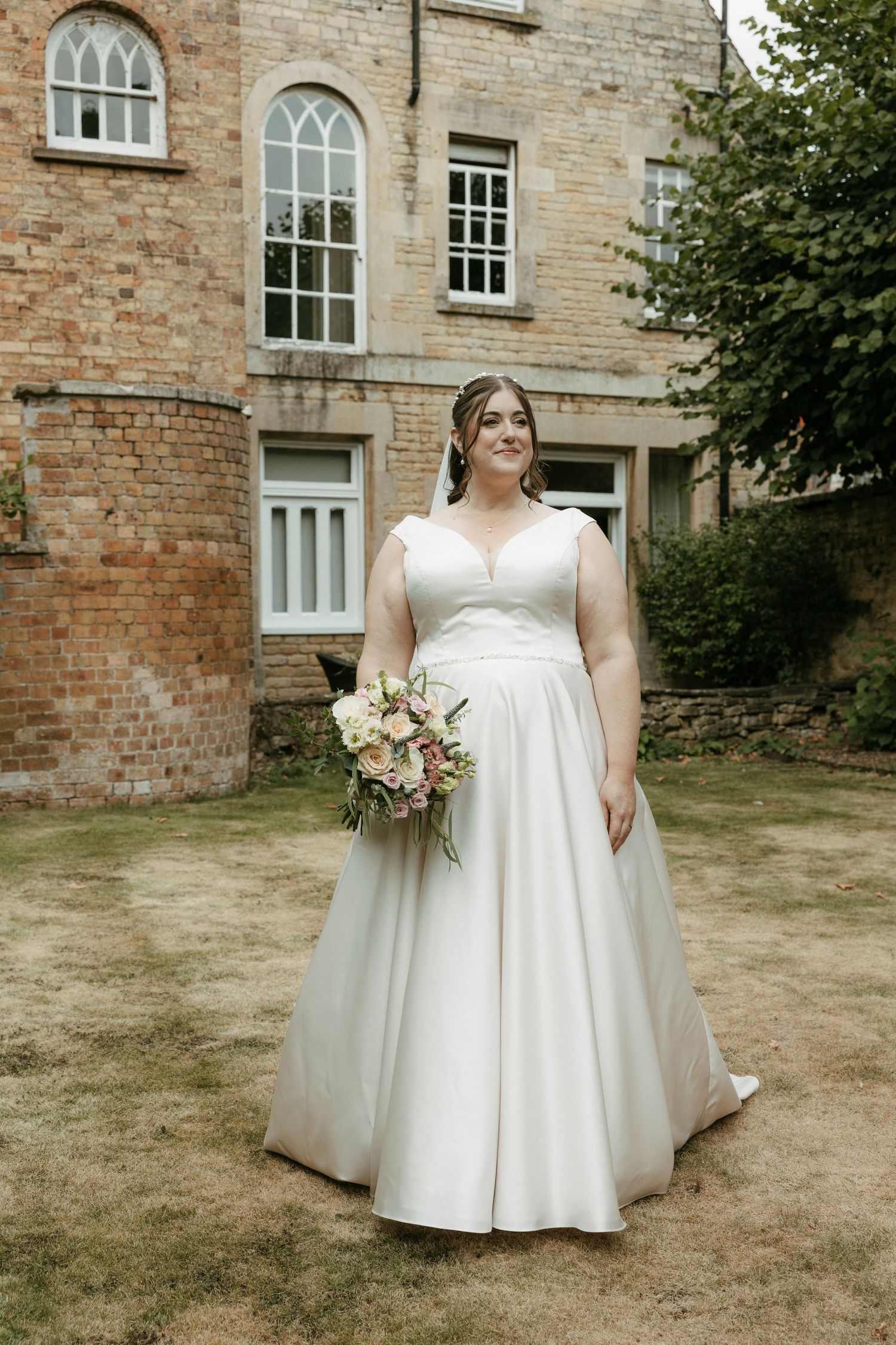 Bride in white satin A-line wedding gown holding bouquet standing on lawn outside historic brick manor house.