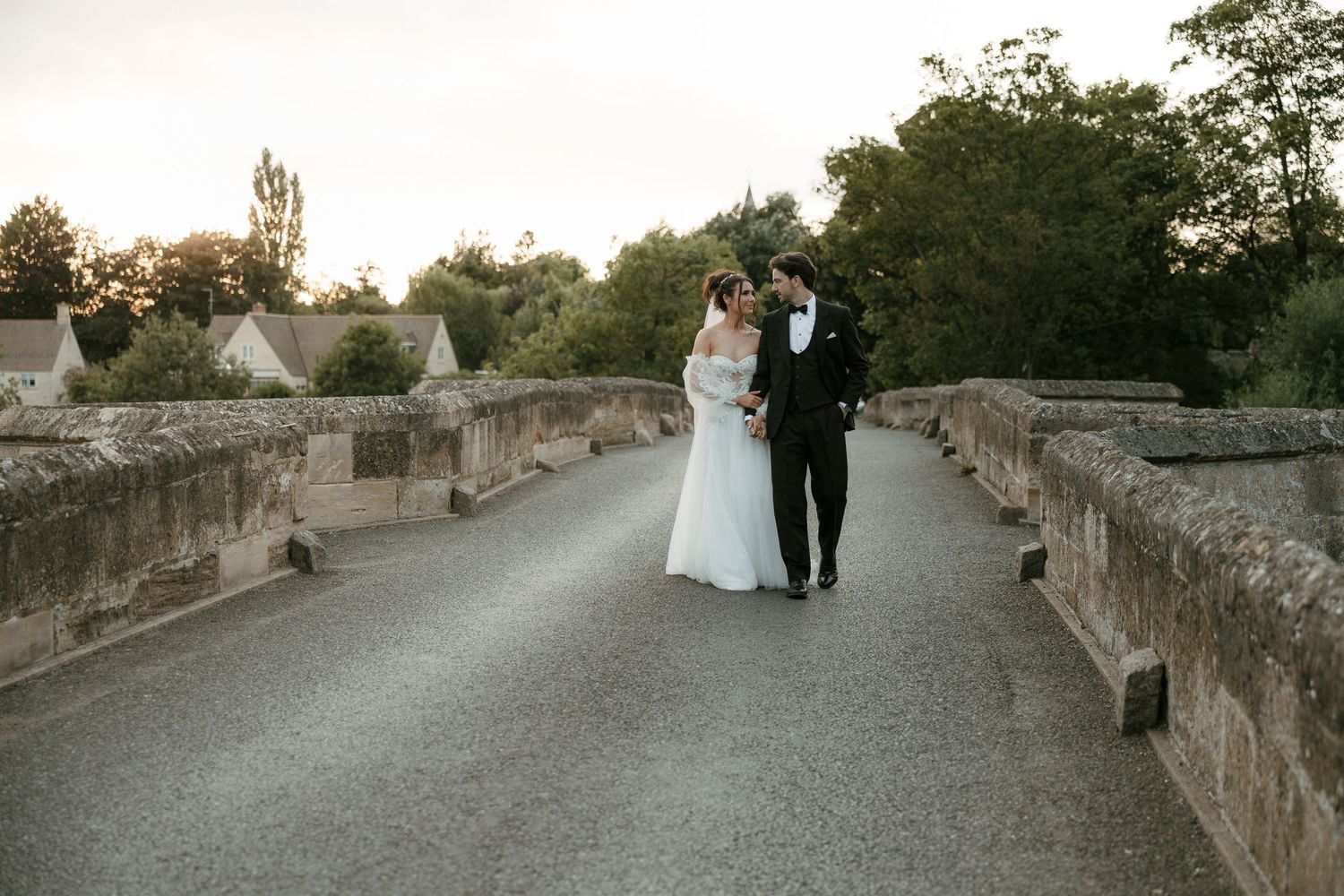 Bride and groom walking hand in hand on a stone bridge at sunset in elegant wedding attire.