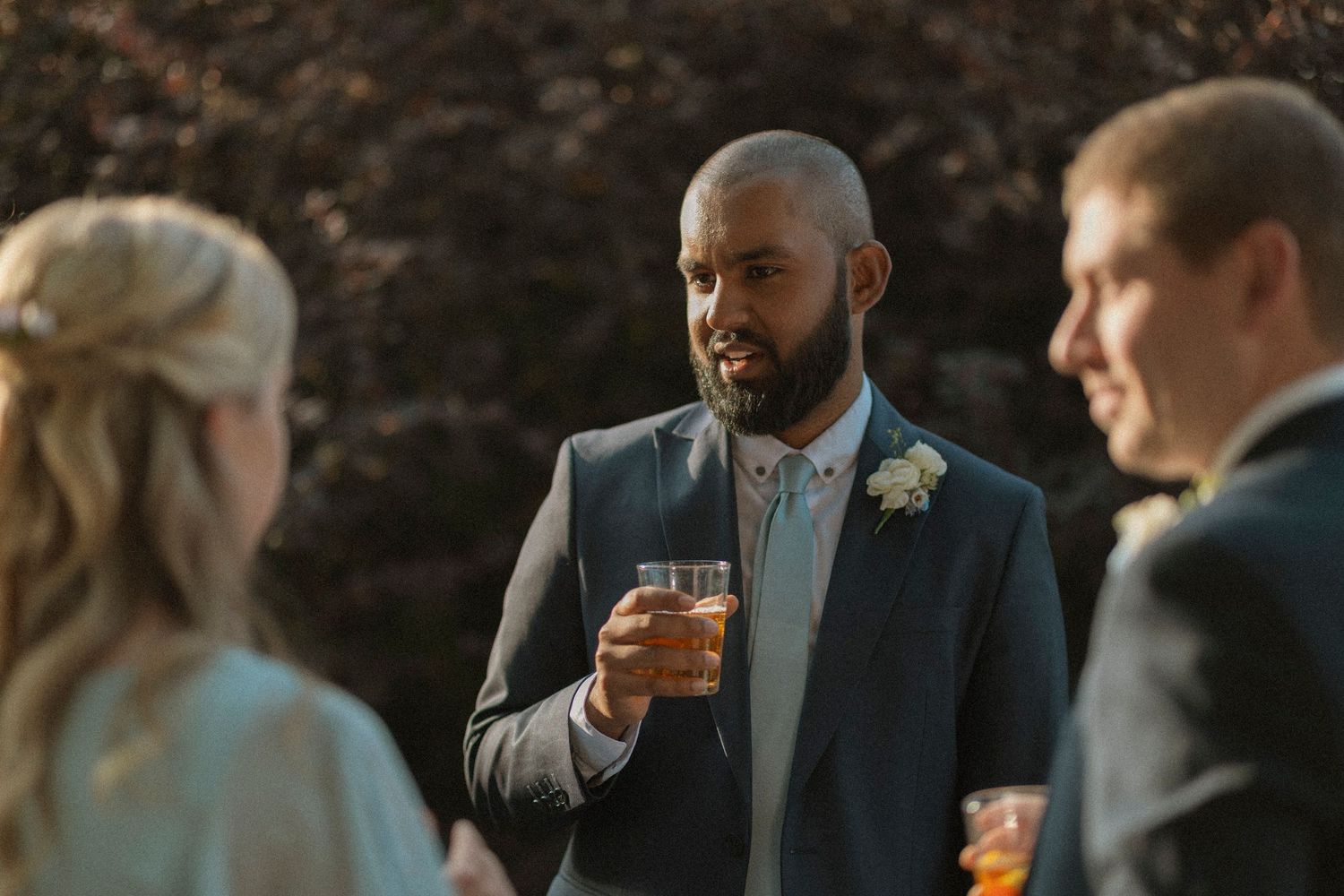 Groom in navy suit holding whiskey glass chatting with wedding guests outdoors at elegant reception.