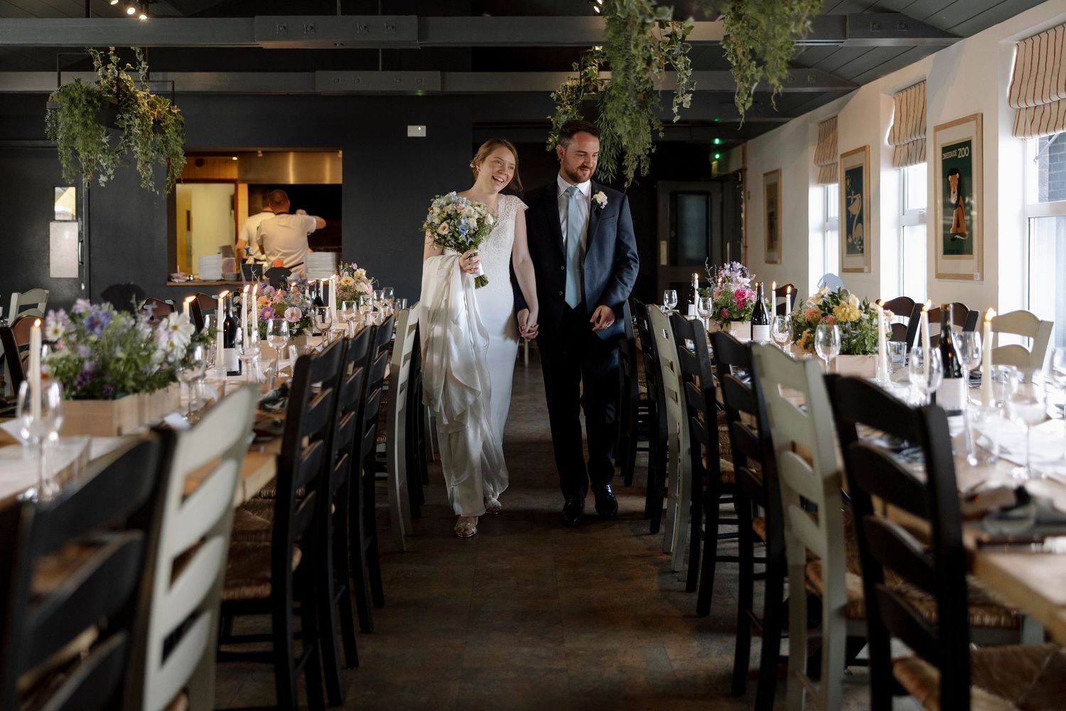 Bride and groom walk hand-in-hand through elegantly decorated wedding reception hall with floral arrangements.
