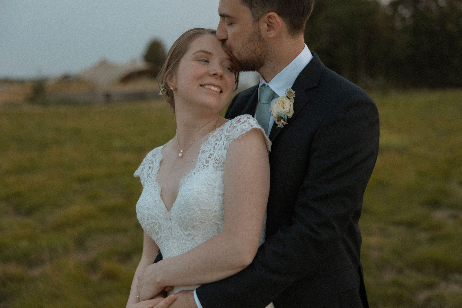 A groom in a navy suit kisses his smiling bride in a lace wedding dress outdoors in a green field.