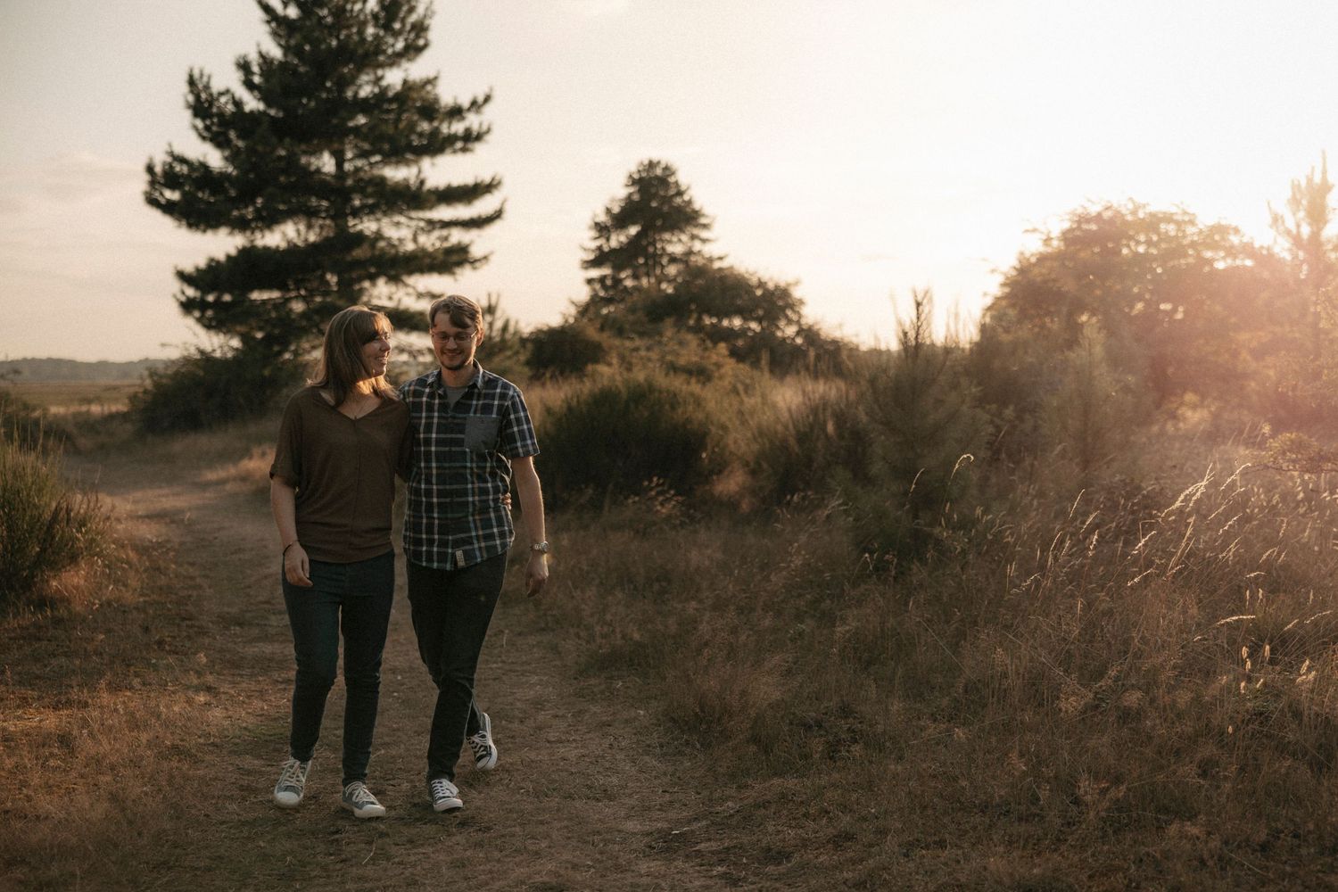 Couple walking hand in hand on a scenic dirt path at golden hour with trees and tall grass in background.