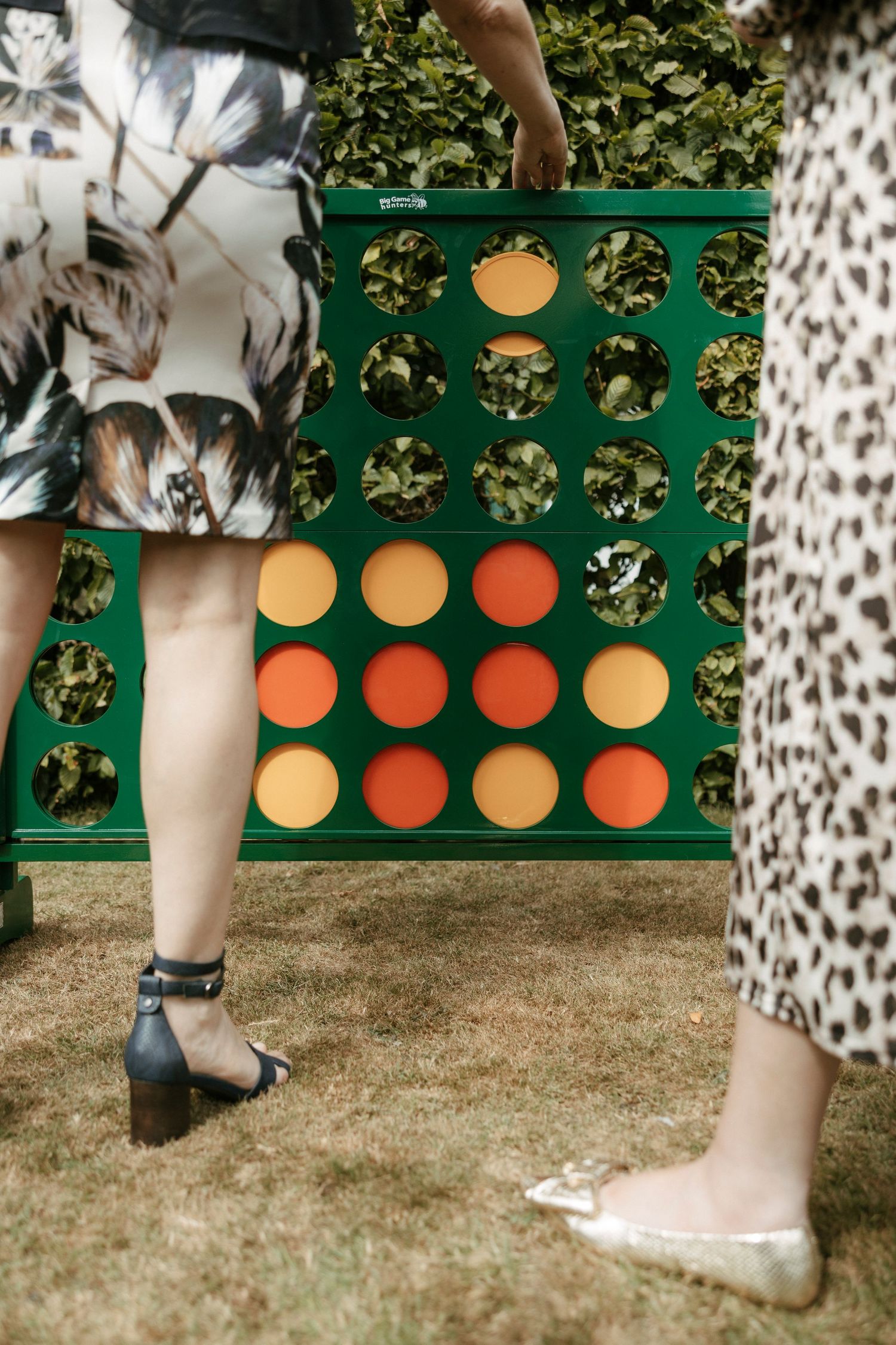 Two wedding guests playing giant Connect Four lawn game on grass at an outdoor garden wedding reception.