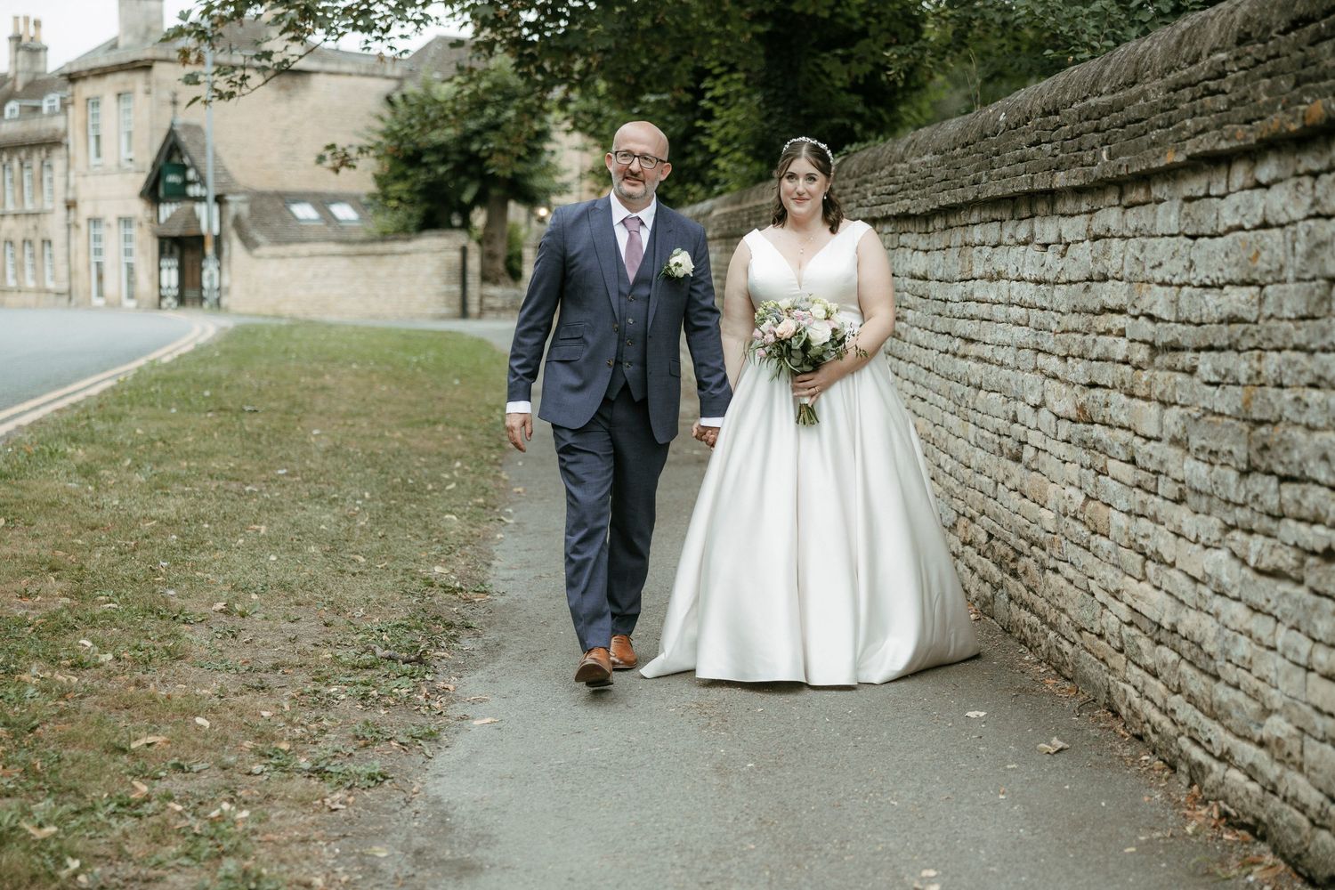Bride and groom walking hand in hand along a stone wall path on their wedding day in a charming village setting.