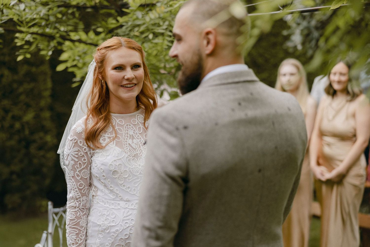Bride in white lace gown smiling at groom during outdoor wedding ceremony with bridesmaids in background.