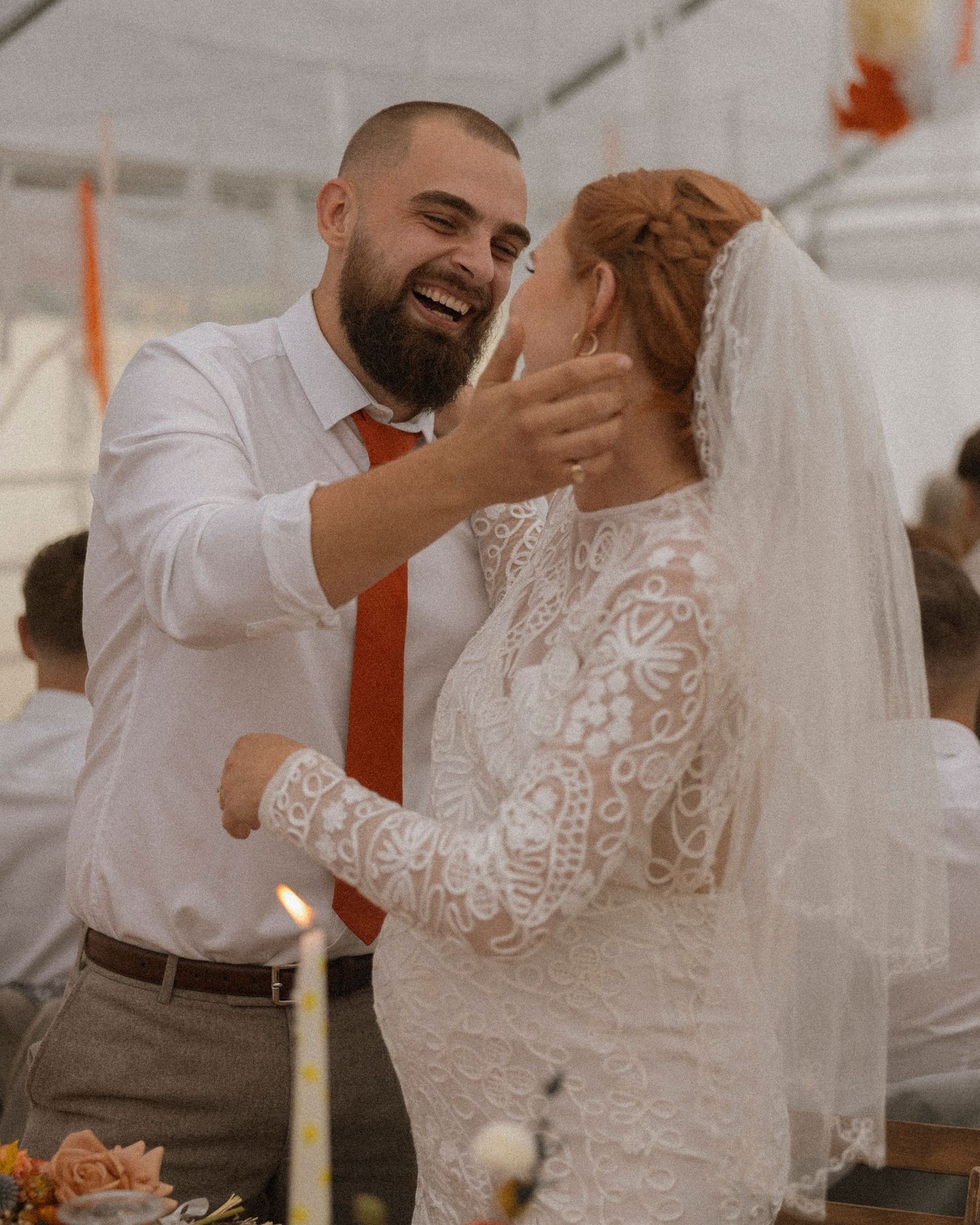 A groom in a white shirt and orange tie tenderly cups his bride's face as they share a joyful moment at their wedding reception.