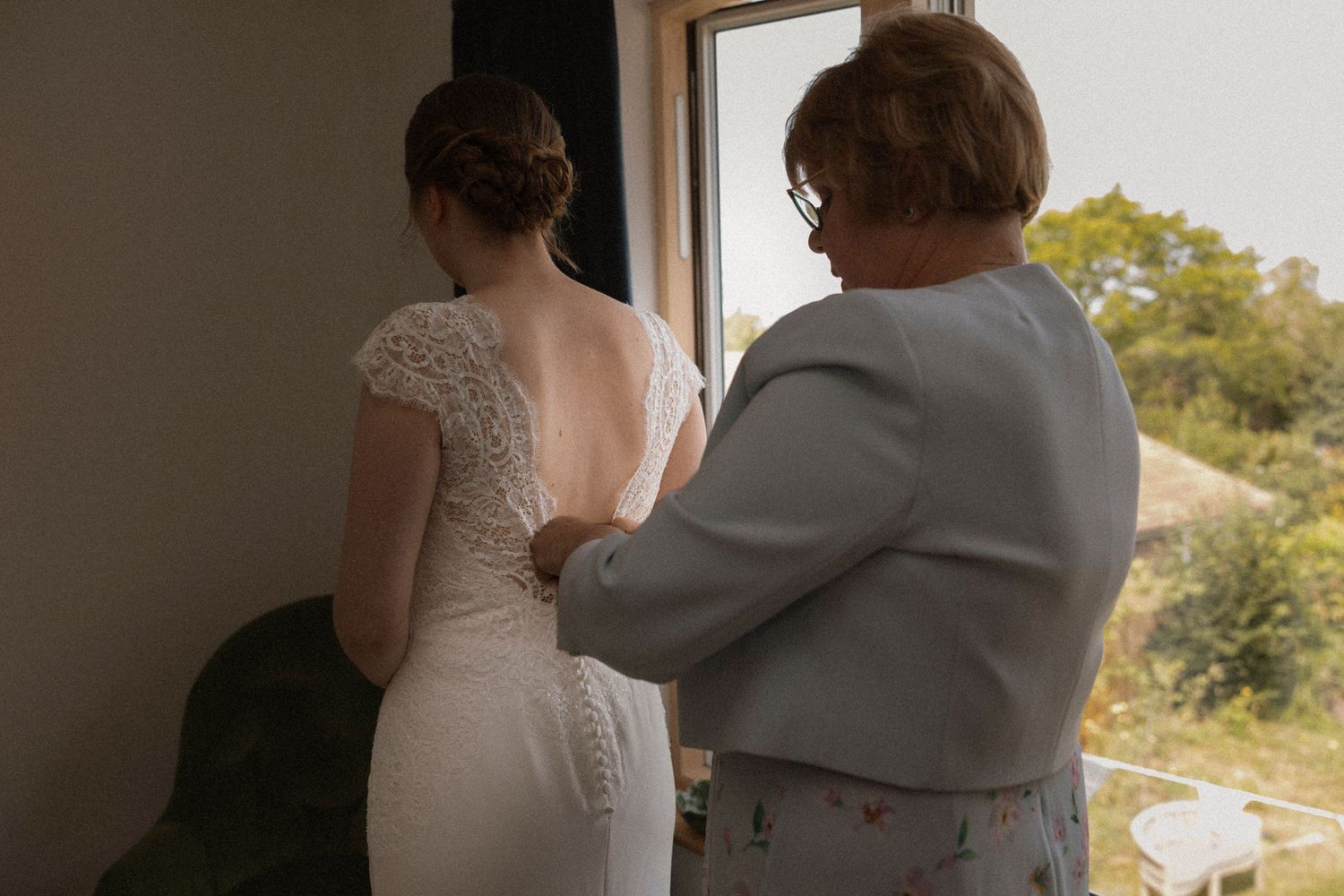 A bride in a white lace wedding dress stands by a window as an older woman helps button the back of her gown.