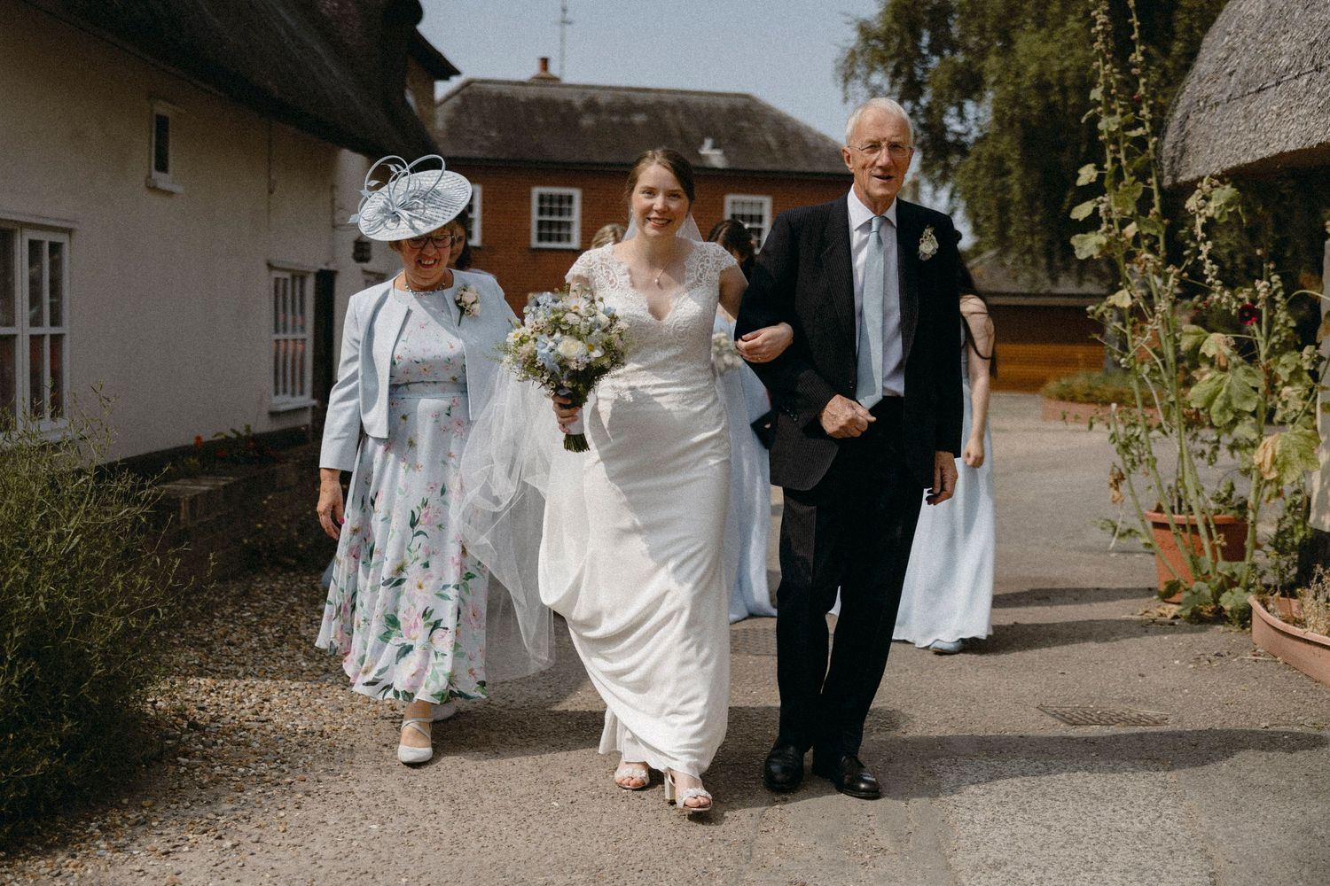 Bride in white gown walks with parents toward wedding venue on sunny day, mother in floral dress and father in suit.