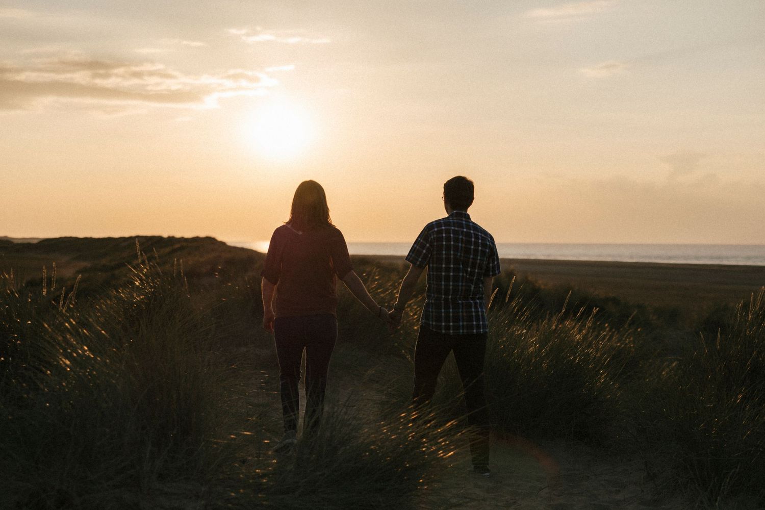 Couple walking hand in hand toward a stunning ocean sunset on a sandy path.