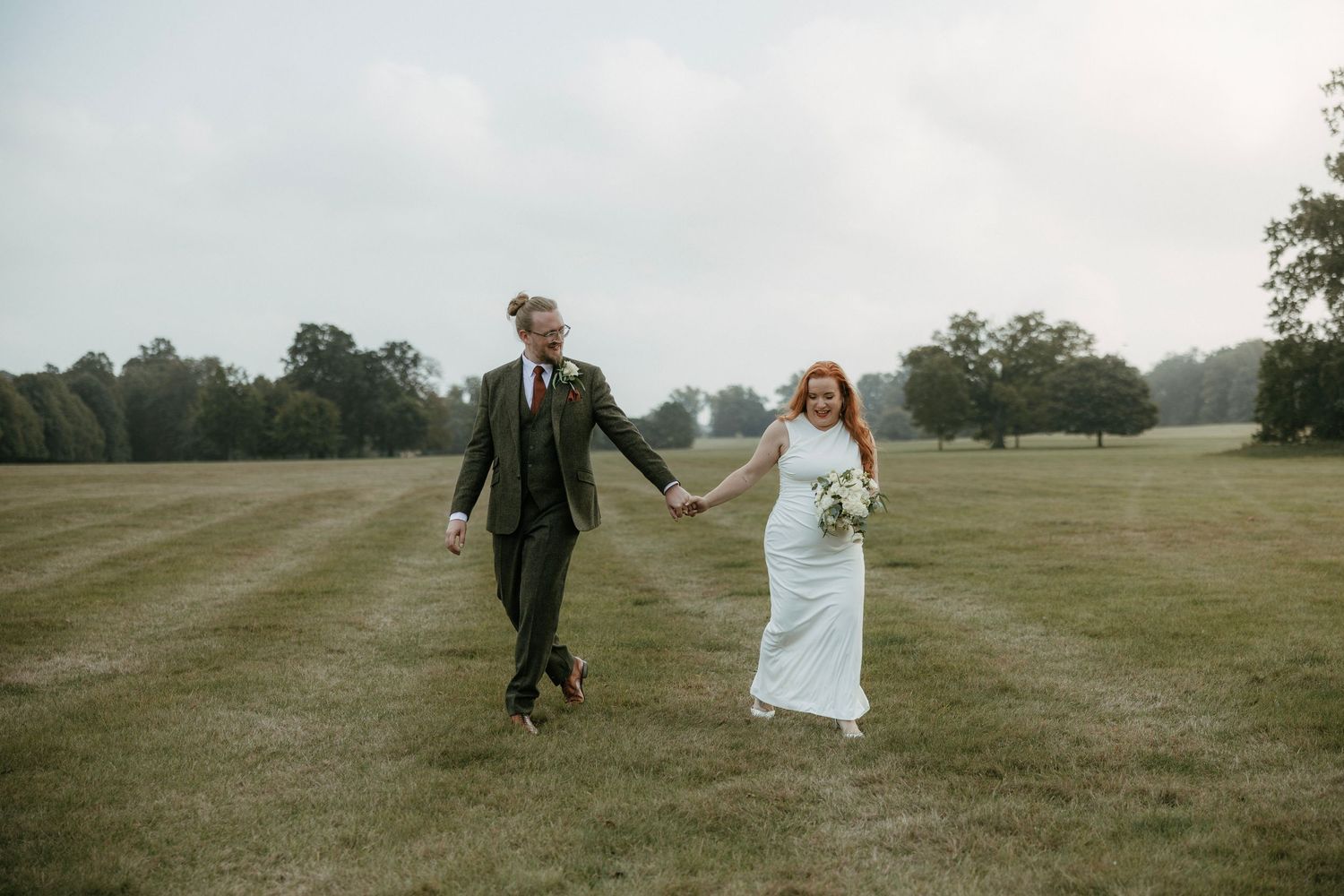 Bride and groom holding hands walking across lush green field on their wedding day in countryside setting.