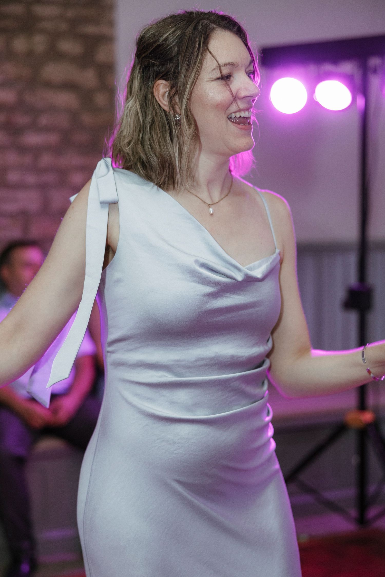 Happy woman in white satin dress dancing joyfully at a wedding reception under purple stage lights.