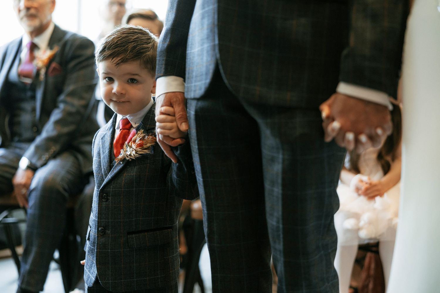 Young ring bearer in navy suit holds adult's hand during wedding ceremony, looking up adorably.