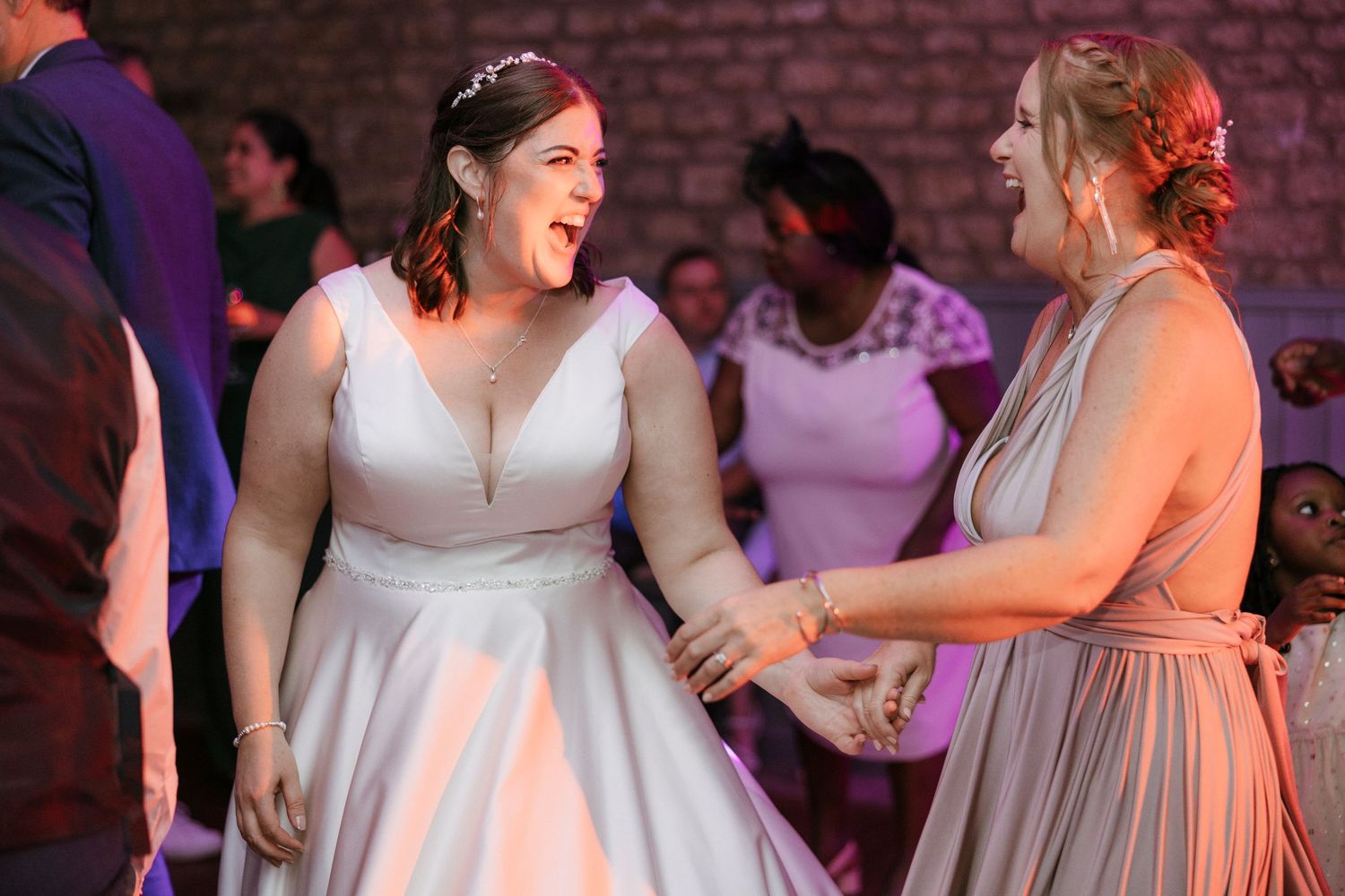 Bride in white gown laughing and dancing with bridesmaid in pink dress at wedding reception.