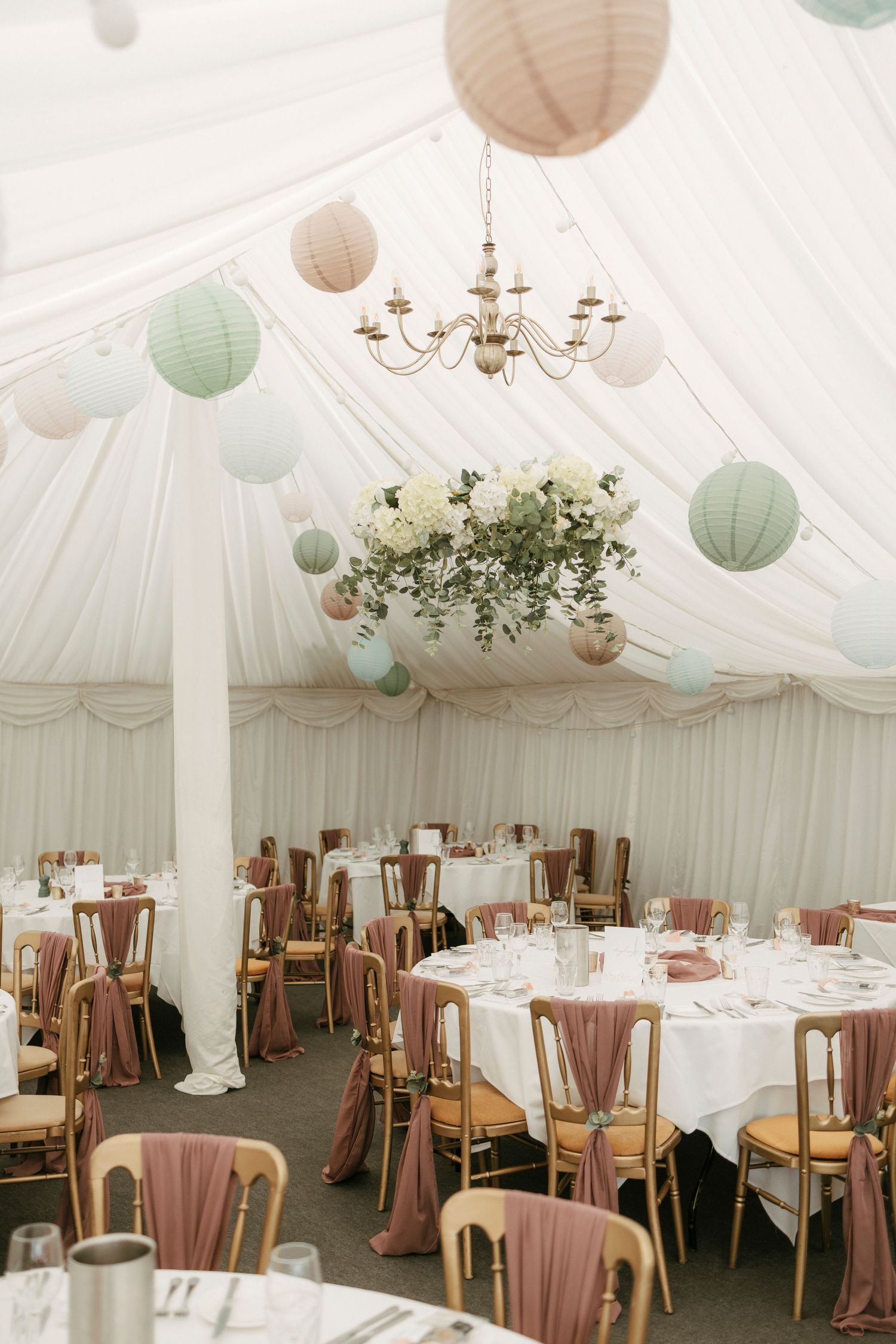 Elegant marquee wedding reception with pastel paper lanterns, chandelier, and dusty rose chair sashes.