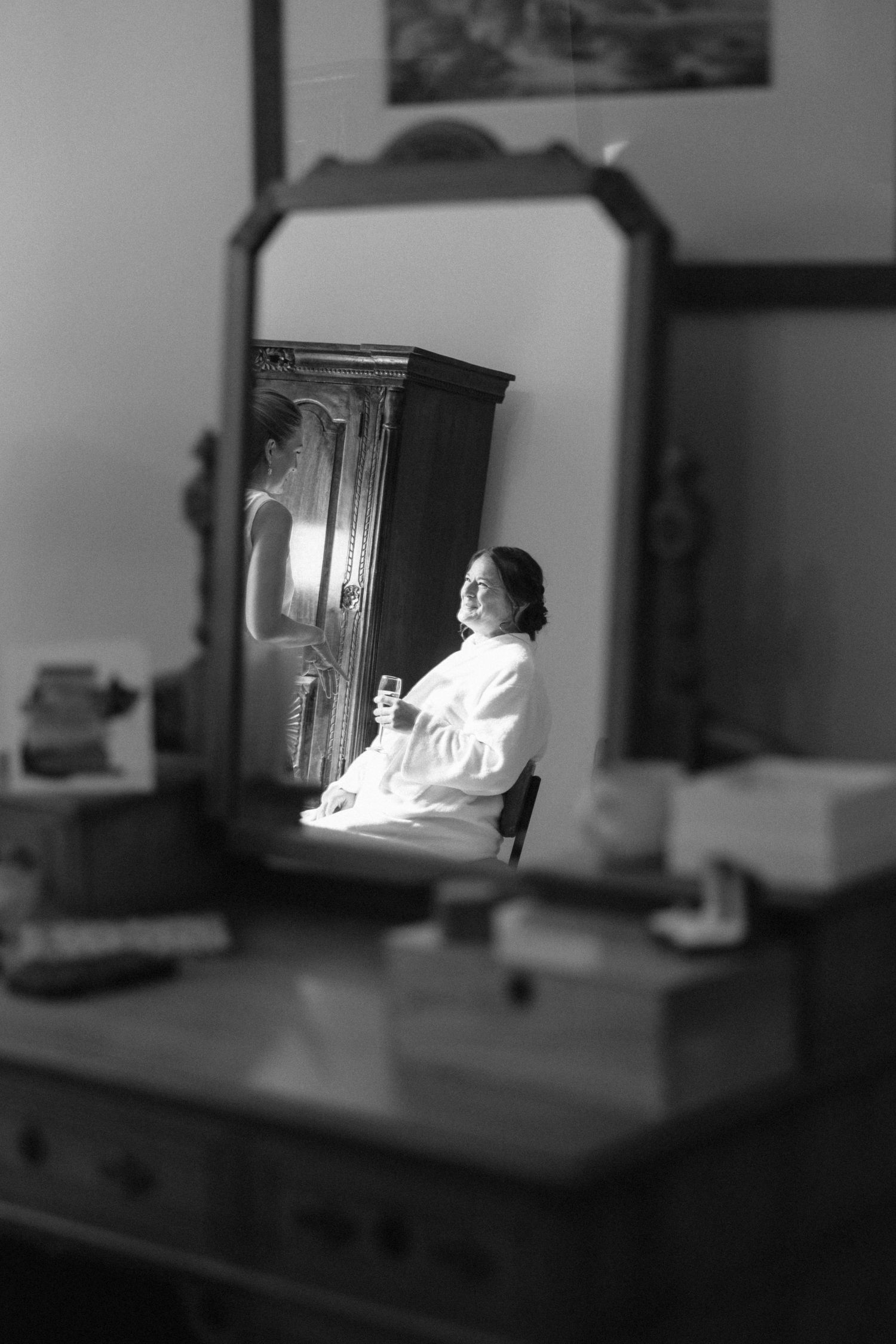 Bride in white robe smiling and holding champagne glass reflected in vintage dressing table mirror.