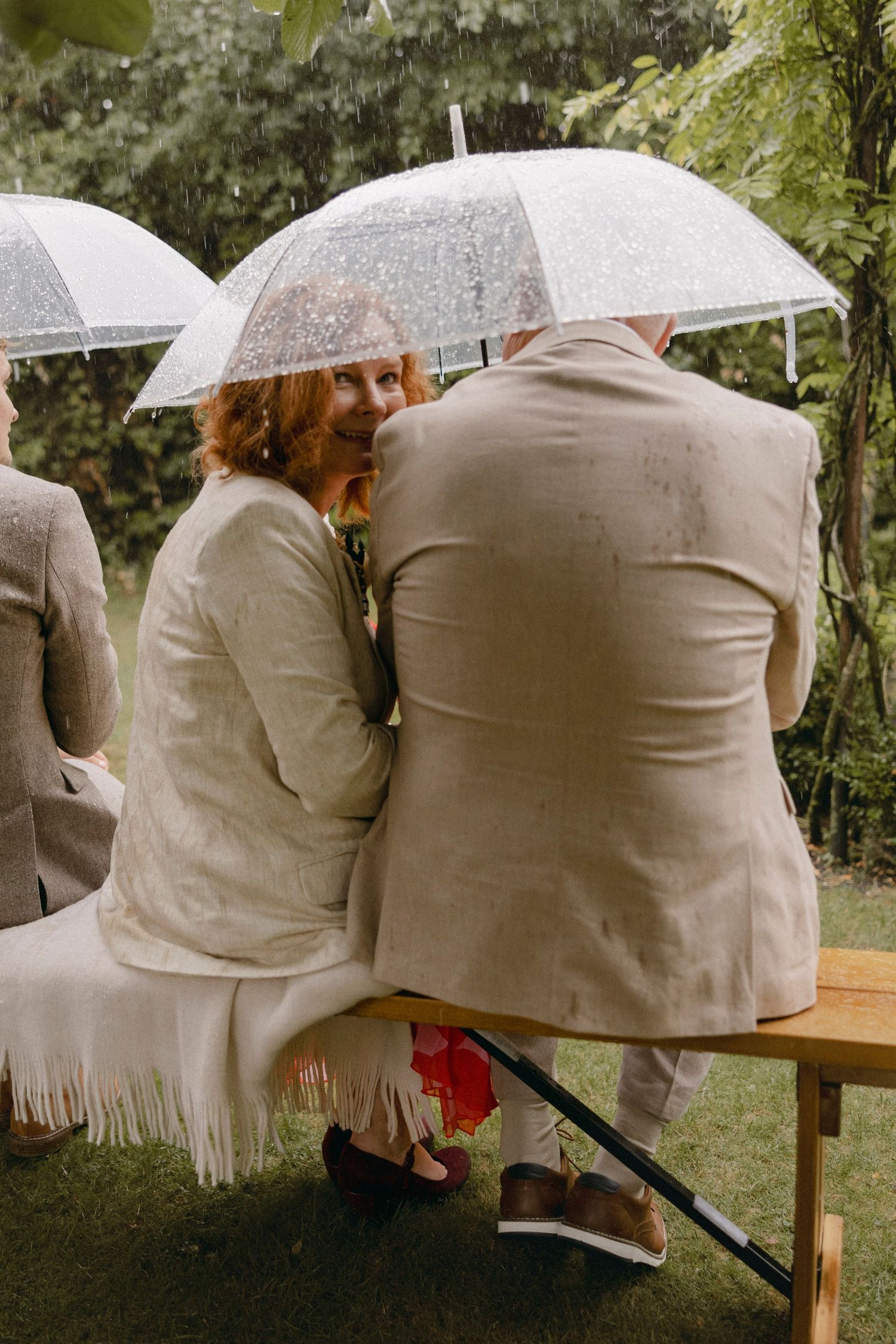 Couple sitting on bench under clear umbrellas in rain at outdoor wedding ceremony surrounded by lush greenery.