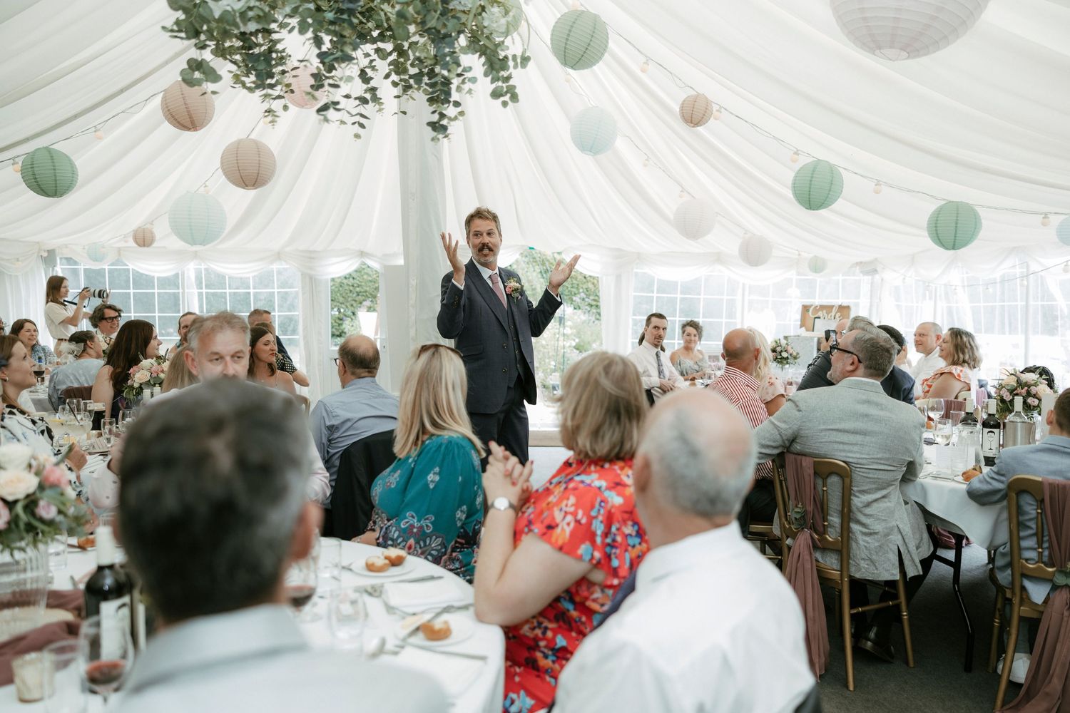 Best man delivering a speech at a wedding reception inside a beautifully decorated marquee with paper lanterns.