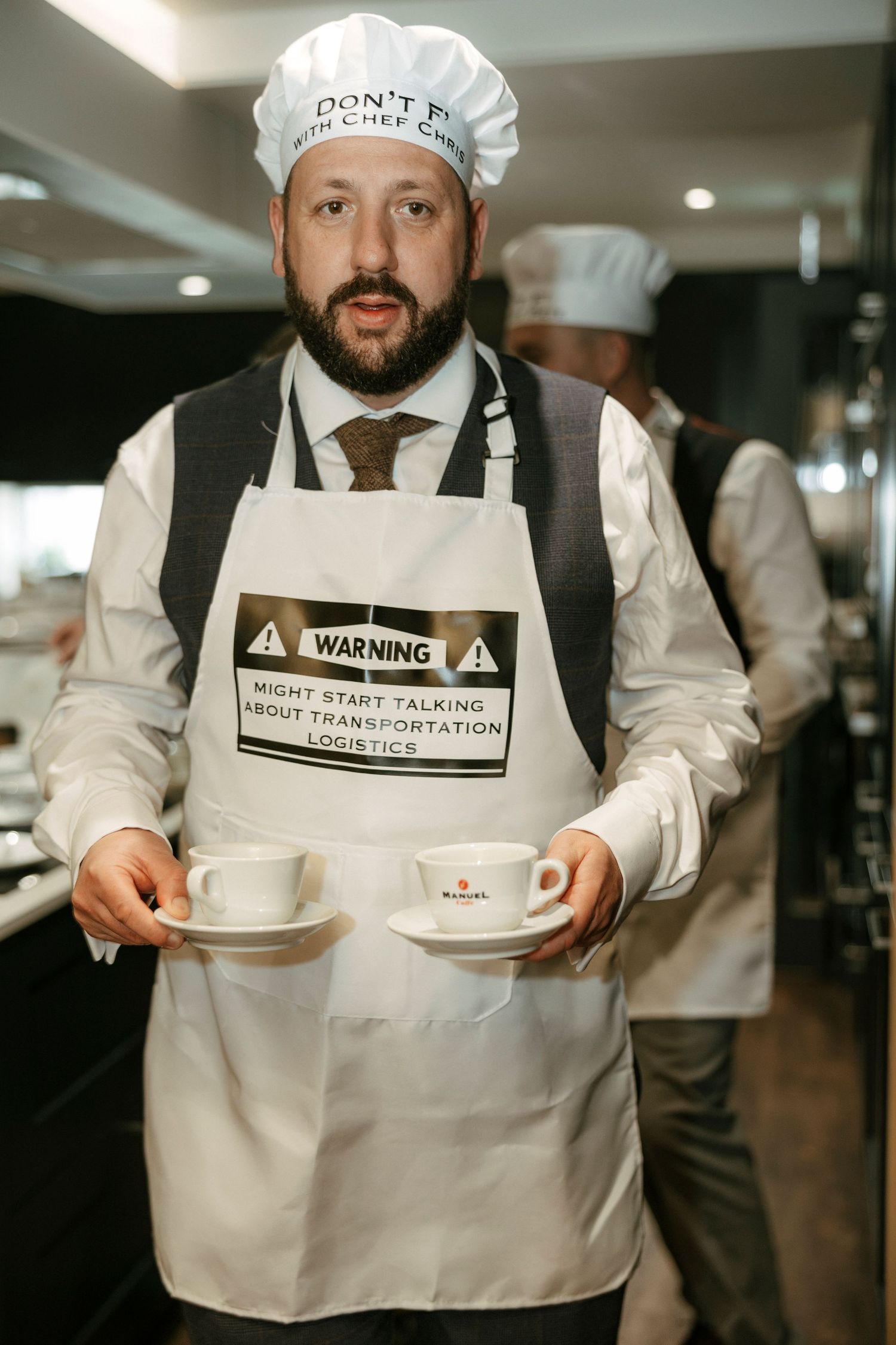 Bearded man in chef hat and warning apron holds two coffee cups in a restaurant kitchen.