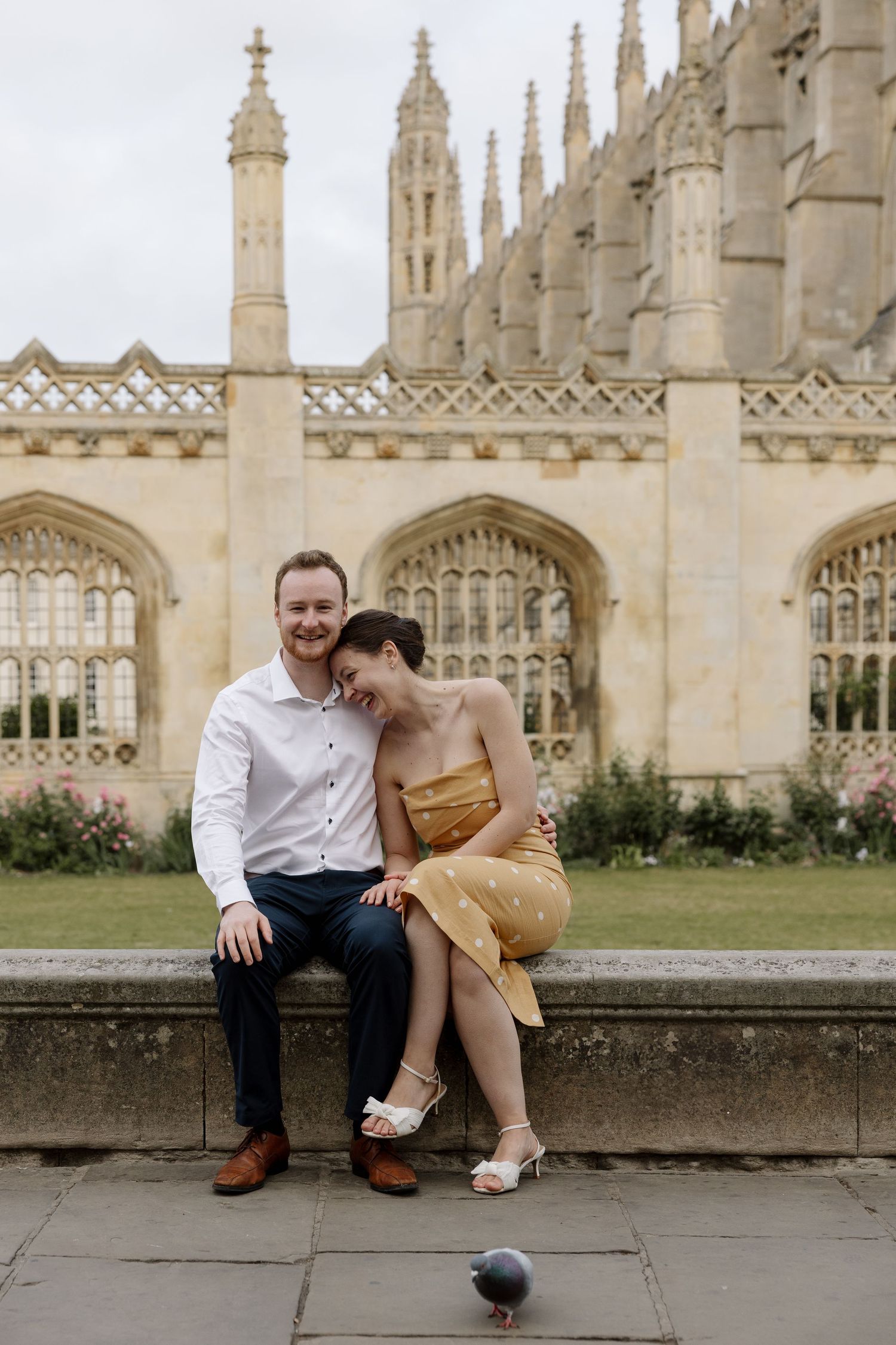 Couple sitting on stone wall in front of King's College Chapel, Cambridge, woman in gold dress leaning on man's shoulder.