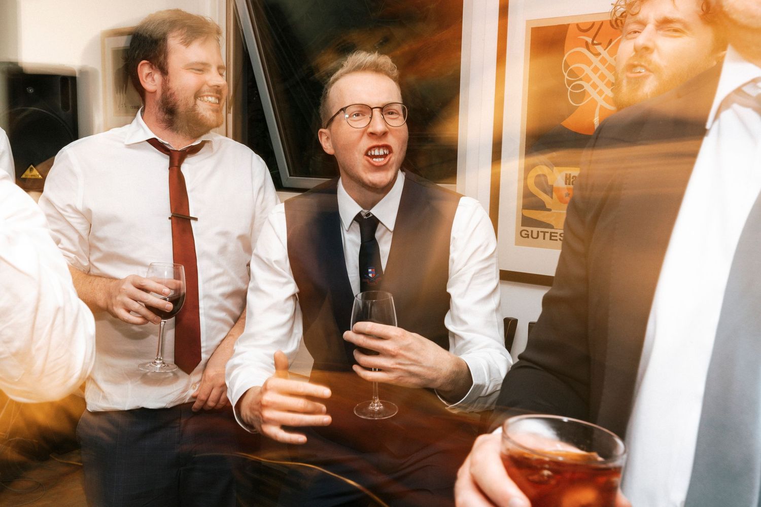Three men in dress shirts and ties laughing and holding drinks at a festive indoor social gathering.