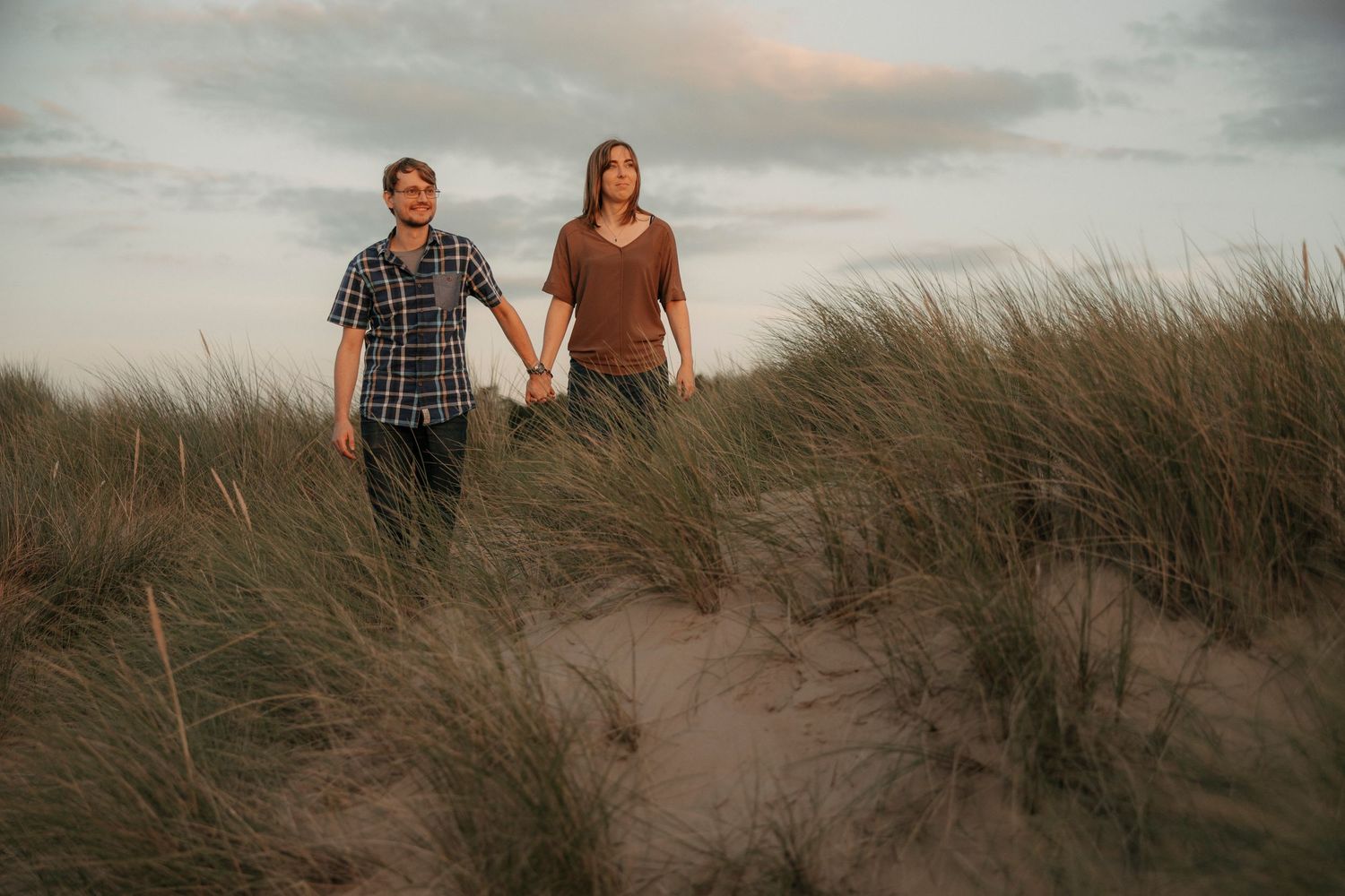 Couple holding hands walking through sandy beach dunes with tall grass under a cloudy sunset sky.