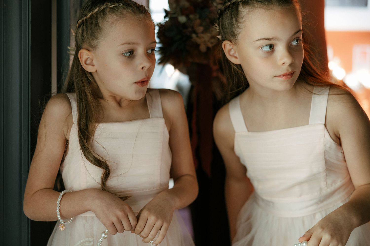 Two young ballerinas in white dresses pose near a window with matching hairstyles and outfits.