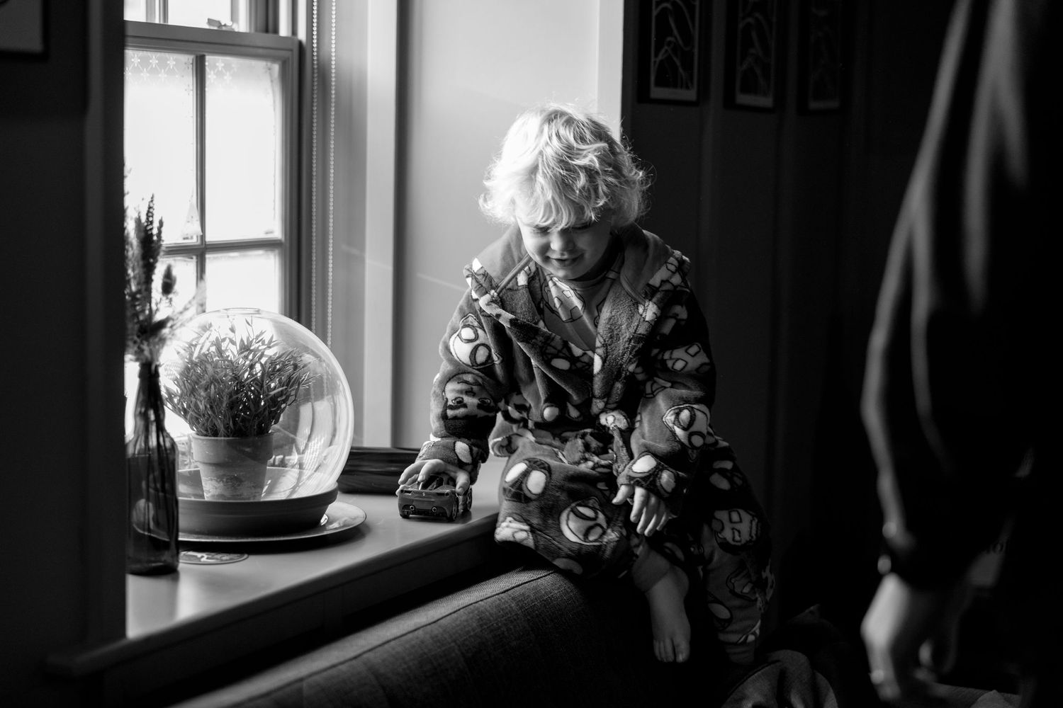 A black and white photograph of someone in a floral robe sitting beside a window with a glass globe.
